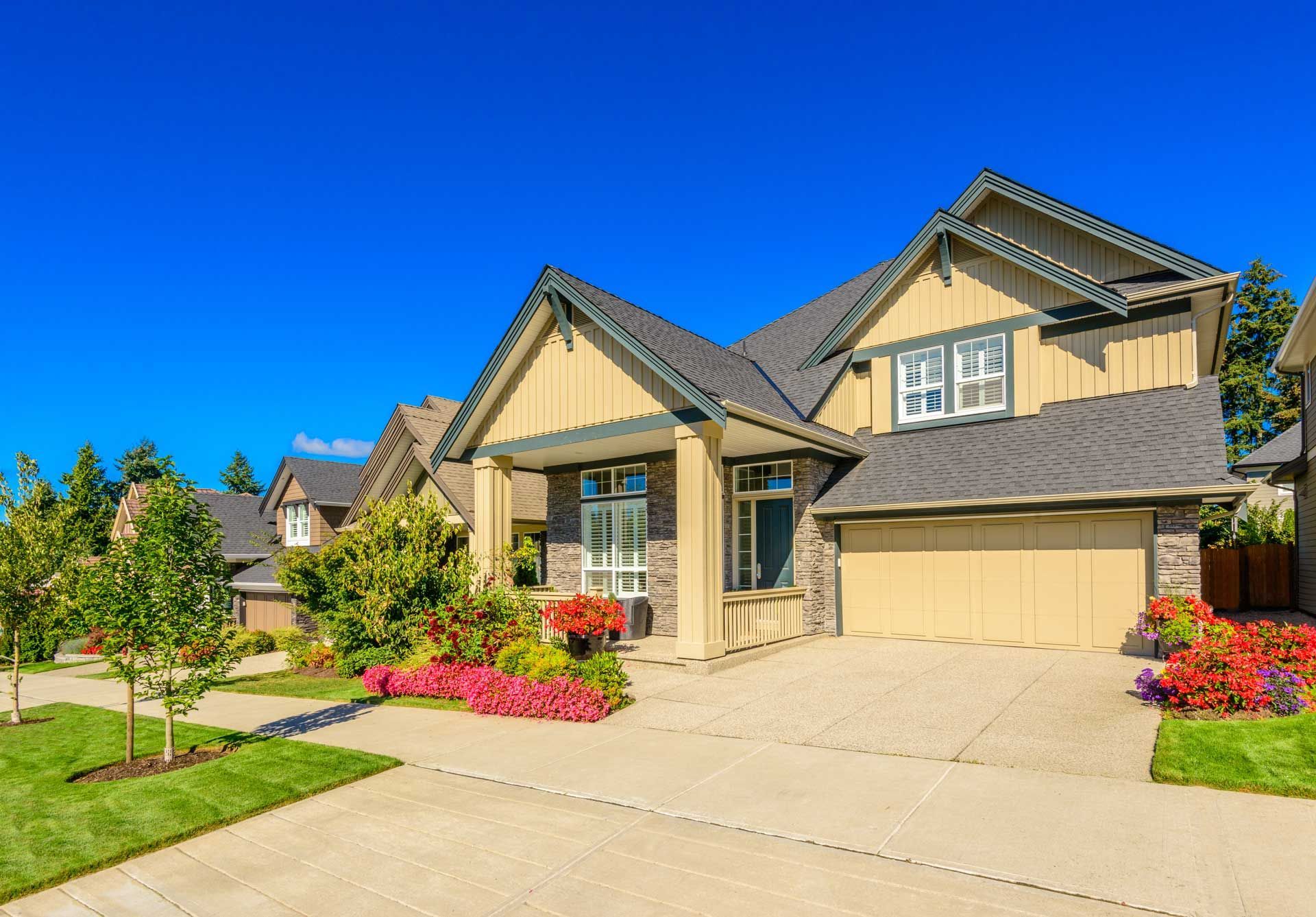 Two-story beige house with a driveway, surrounded by landscaping, under a clear blue sky.