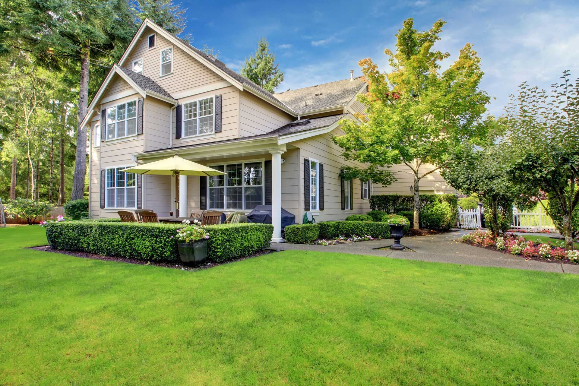 Beige two-story house with green lawn, trees, and patio with umbrella.