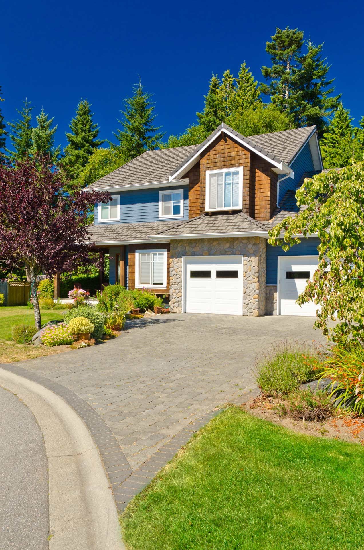 Blue and brown two-story house with a stone and white garage. Paved driveway, green lawn, trees, and blue sky.