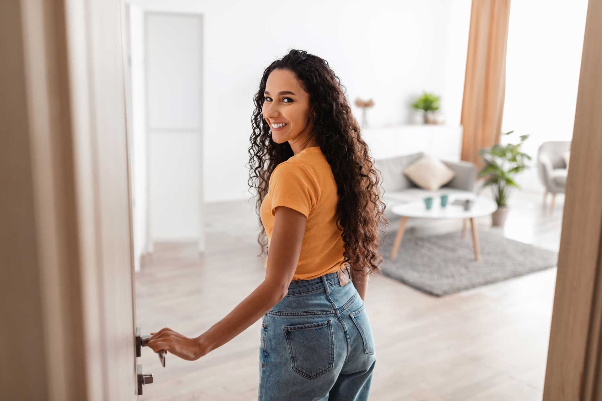 Woman smiles, opens door, looks back. Enters bright living room with gray rug, sofa, and coffee table.