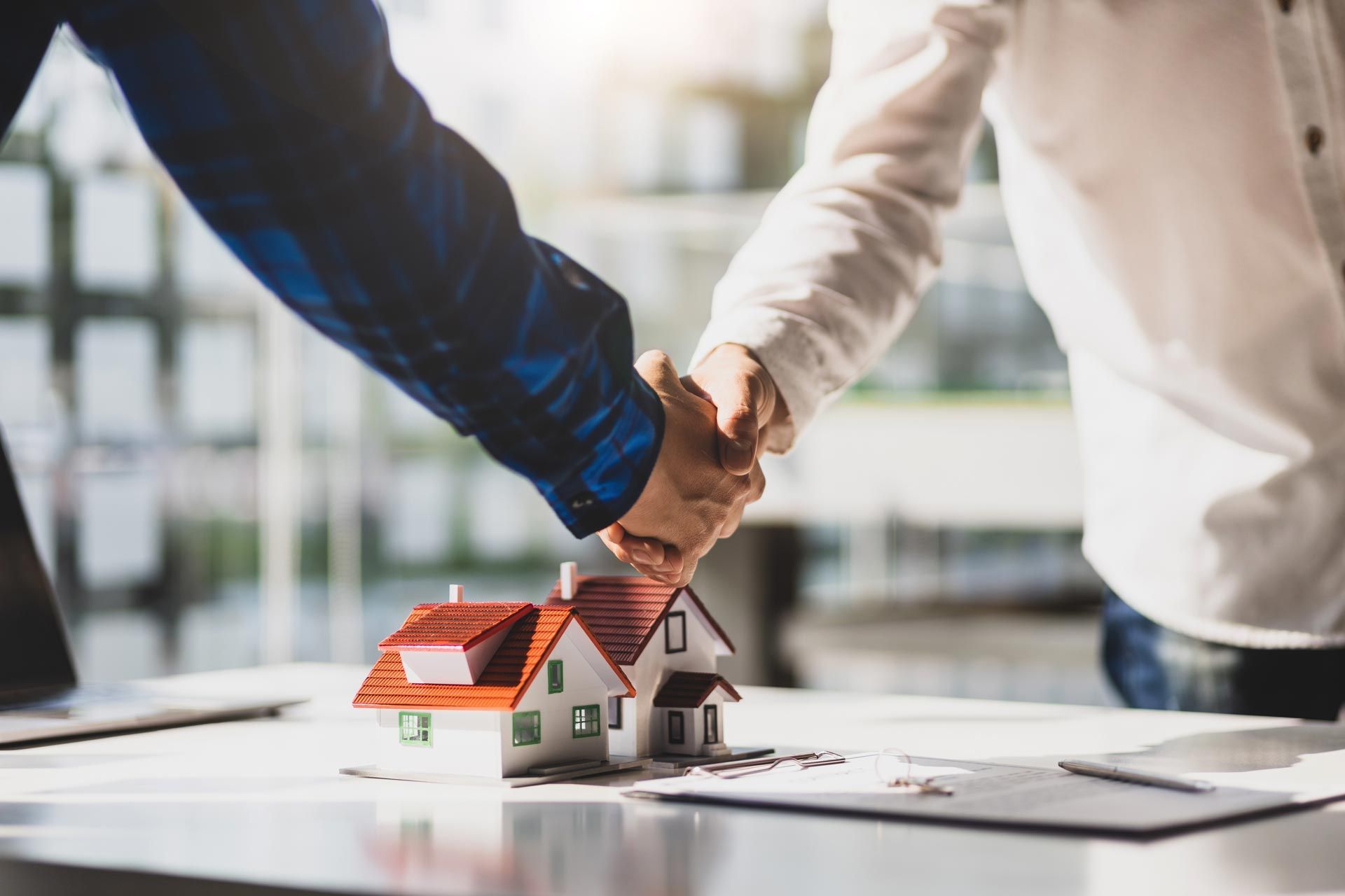 Two people shaking hands over a miniature house, symbolizing a real estate deal.