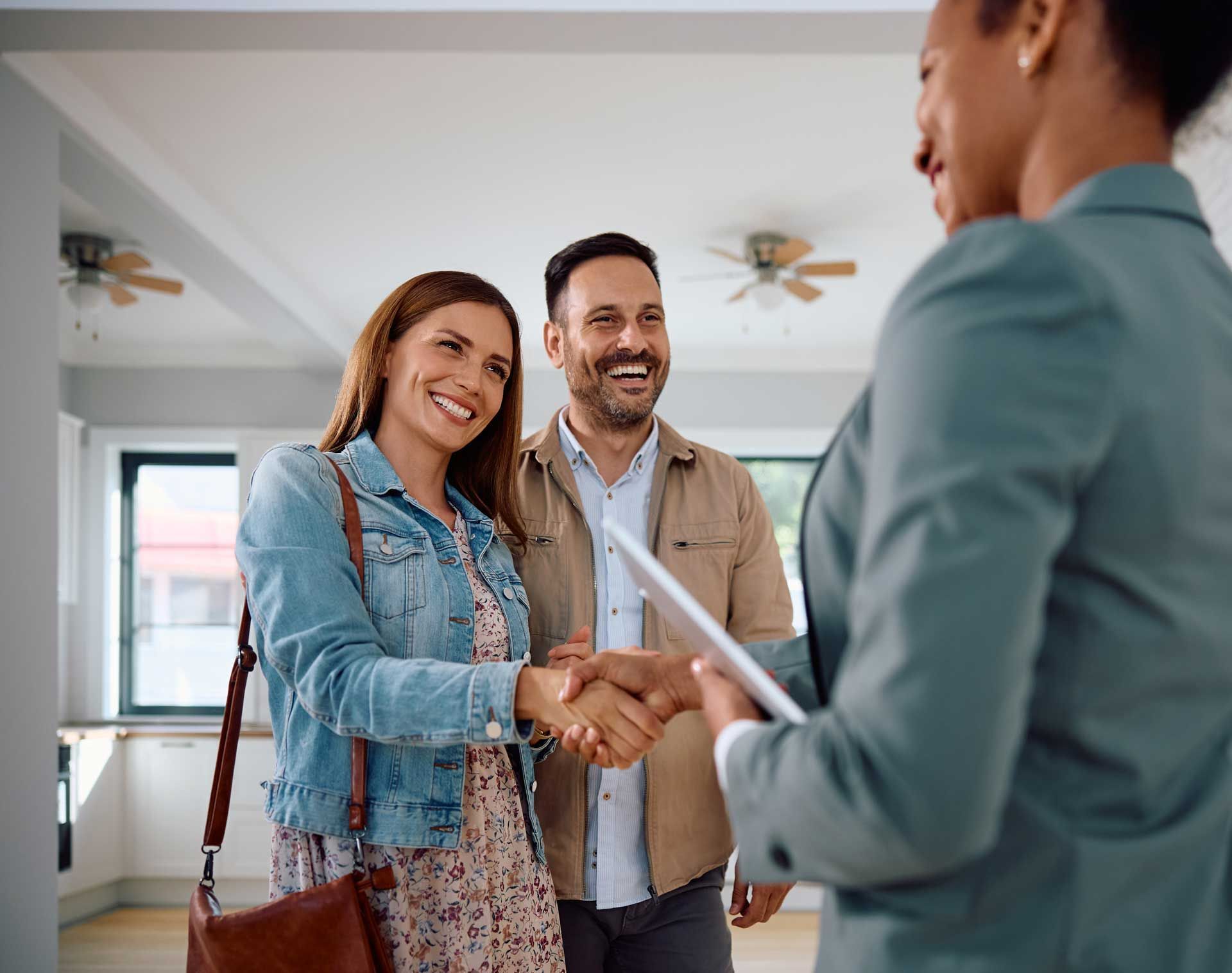 Couple shaking hands with a realtor in an empty room, smiling.