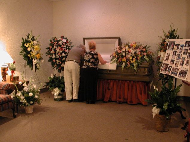 Two people at an open casket in a funeral home, surrounded by floral arrangements and a memorial photo display.