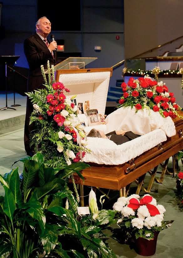 A man speaks near an open casket. Red and white flowers adorn the casket; a speaker is in front.