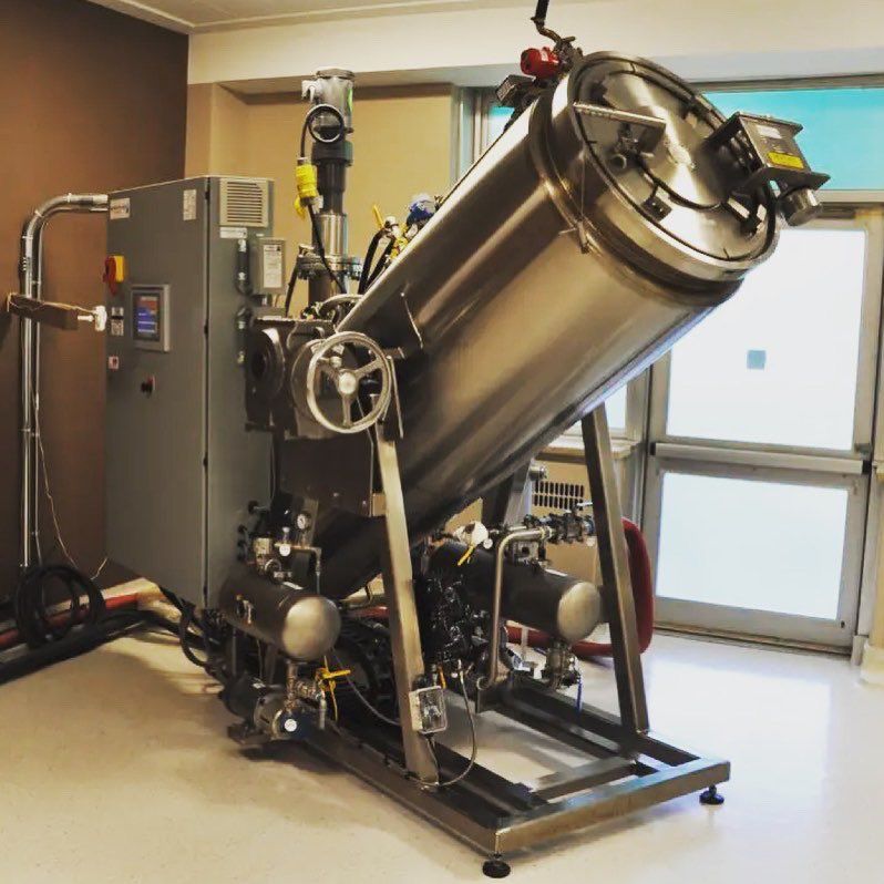 Large, tilted, stainless steel industrial autoclave with control panel in a room with a window.