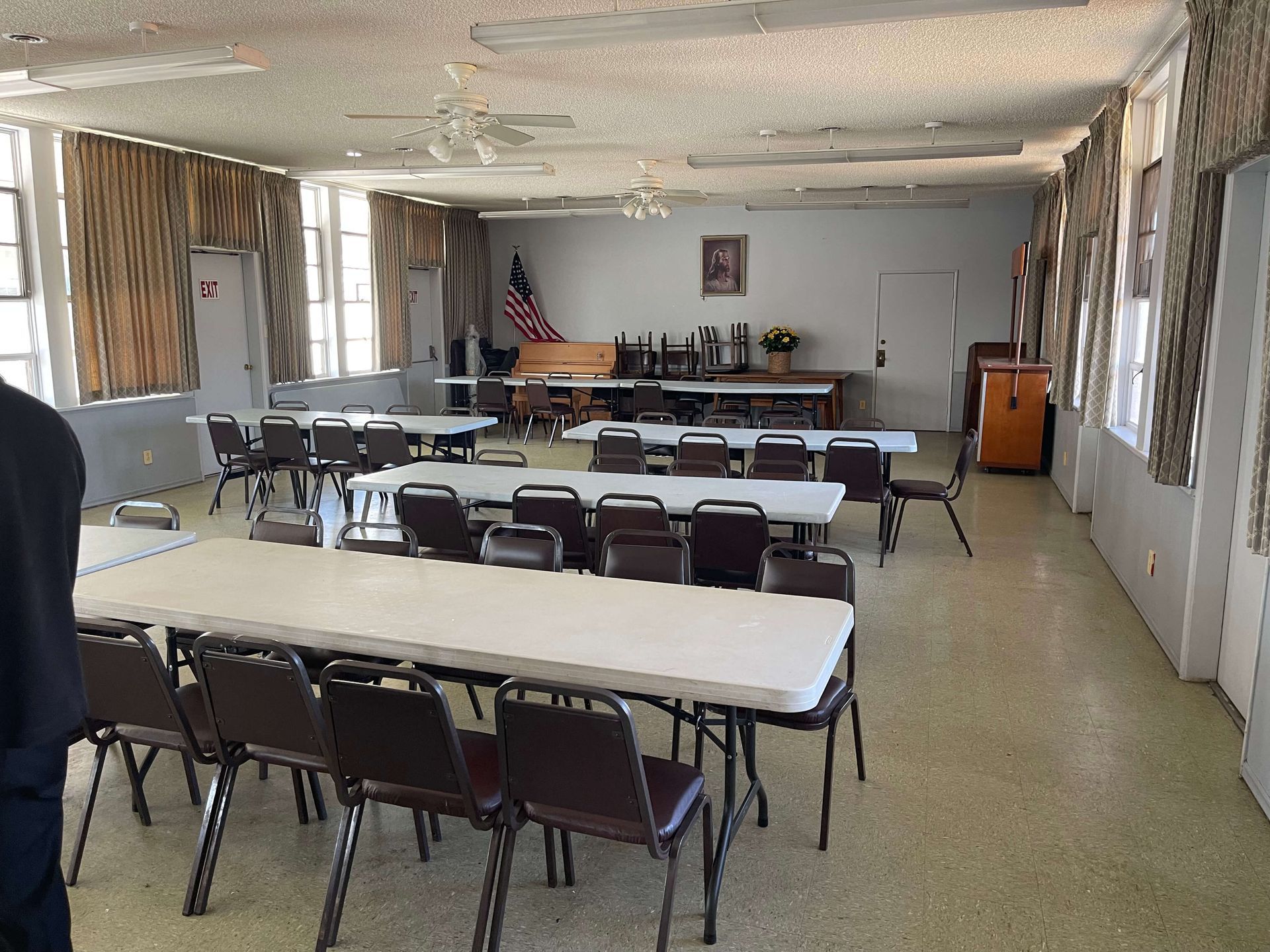 Empty room with rectangular tables and chairs.  An American flag and stage at the back.