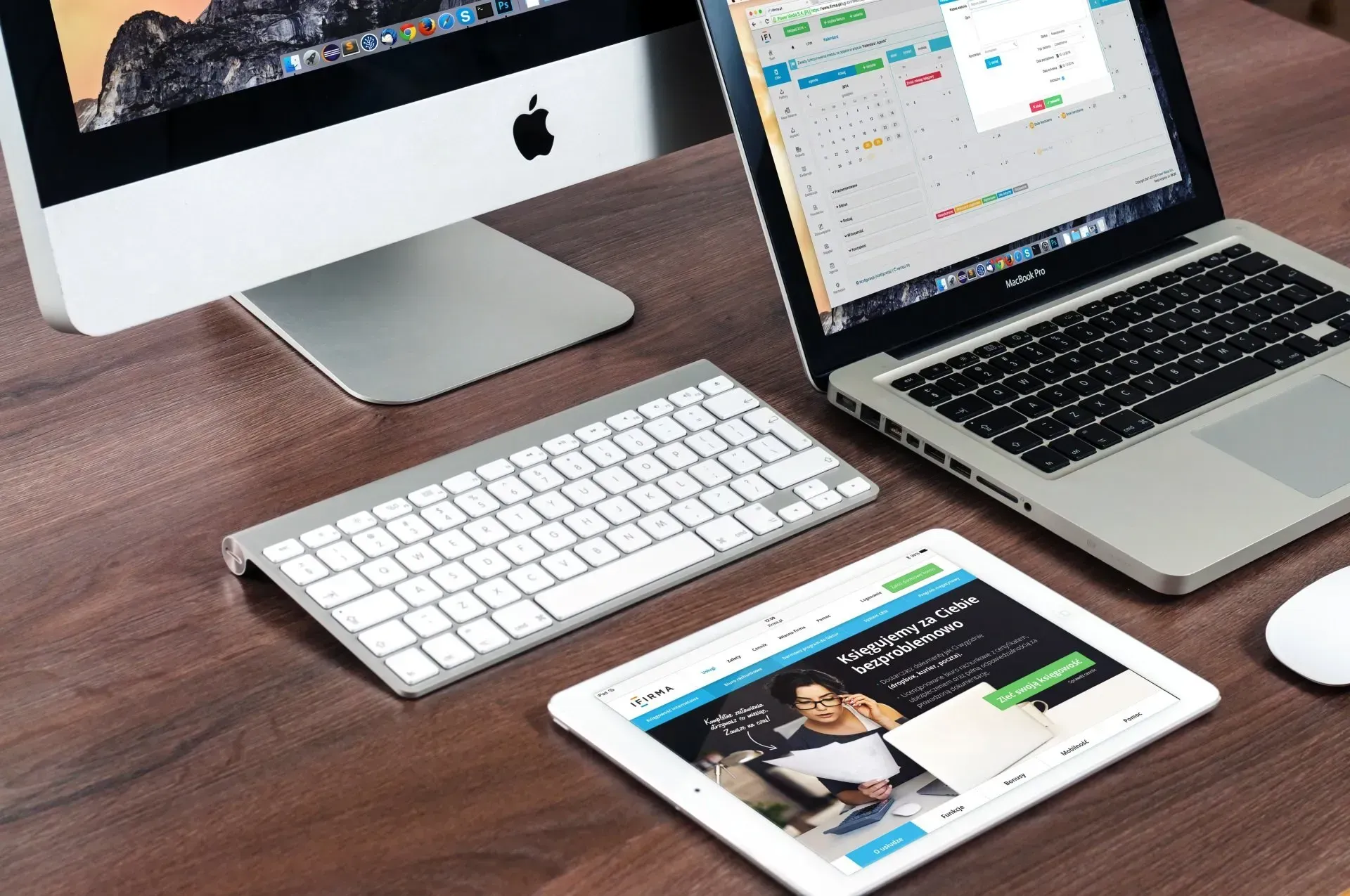 Computer, laptop, tablet, and keyboard on a wooden desk.