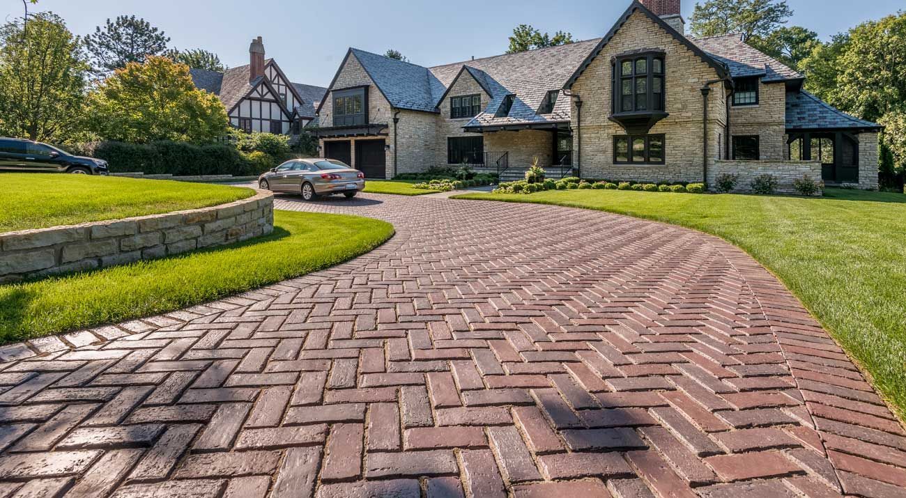 A car is driving down a brick driveway towards a large house.