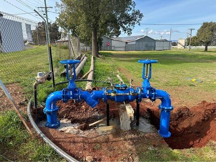 Plumbing pipes and fixtures mounted on a gray cinder block wall. Copper pipes, black pipes, and support brackets. Orange NSW