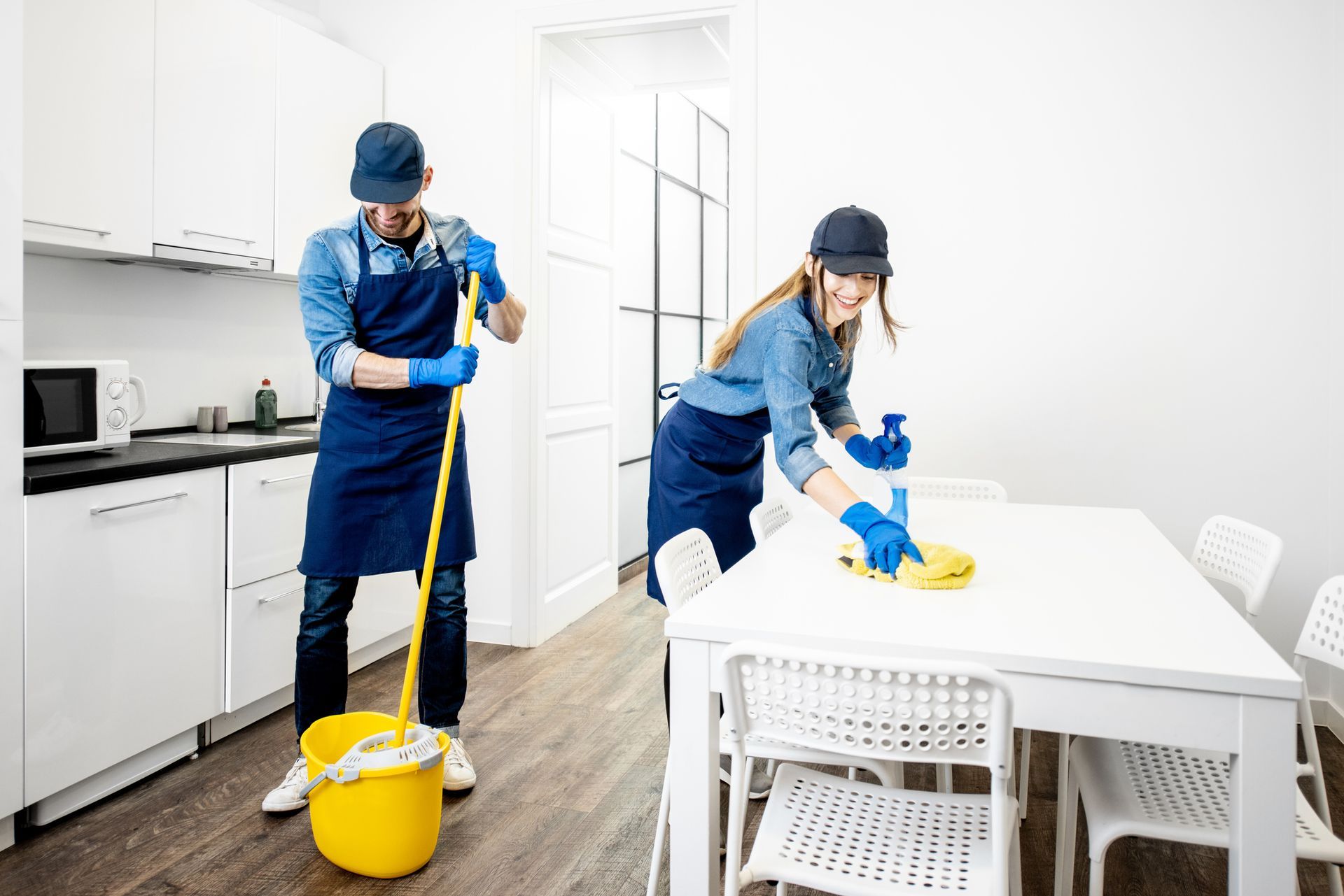 Two people cleaning a white room. One mops the floor, the other wipes a white table with spray.