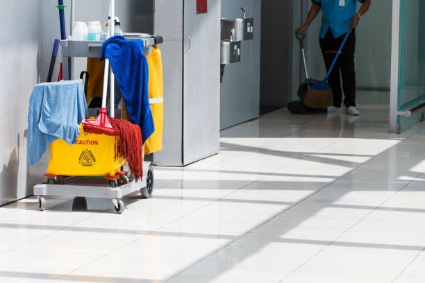 Janitor sweeping a bright, tiled hallway; cleaning cart with supplies to the left.