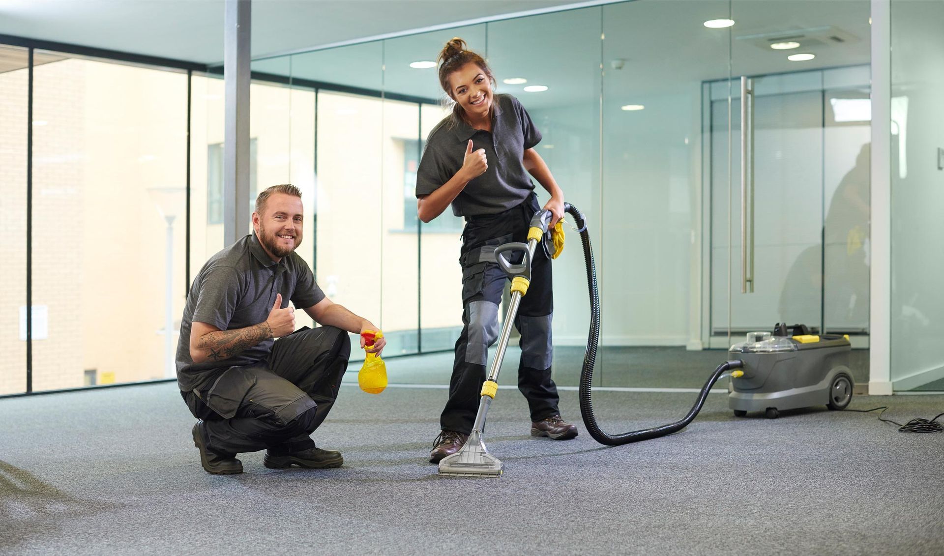 Two people cleaning carpet in an office space. One kneels giving a thumbs up, the other operates a floor cleaning machine, both smiling.