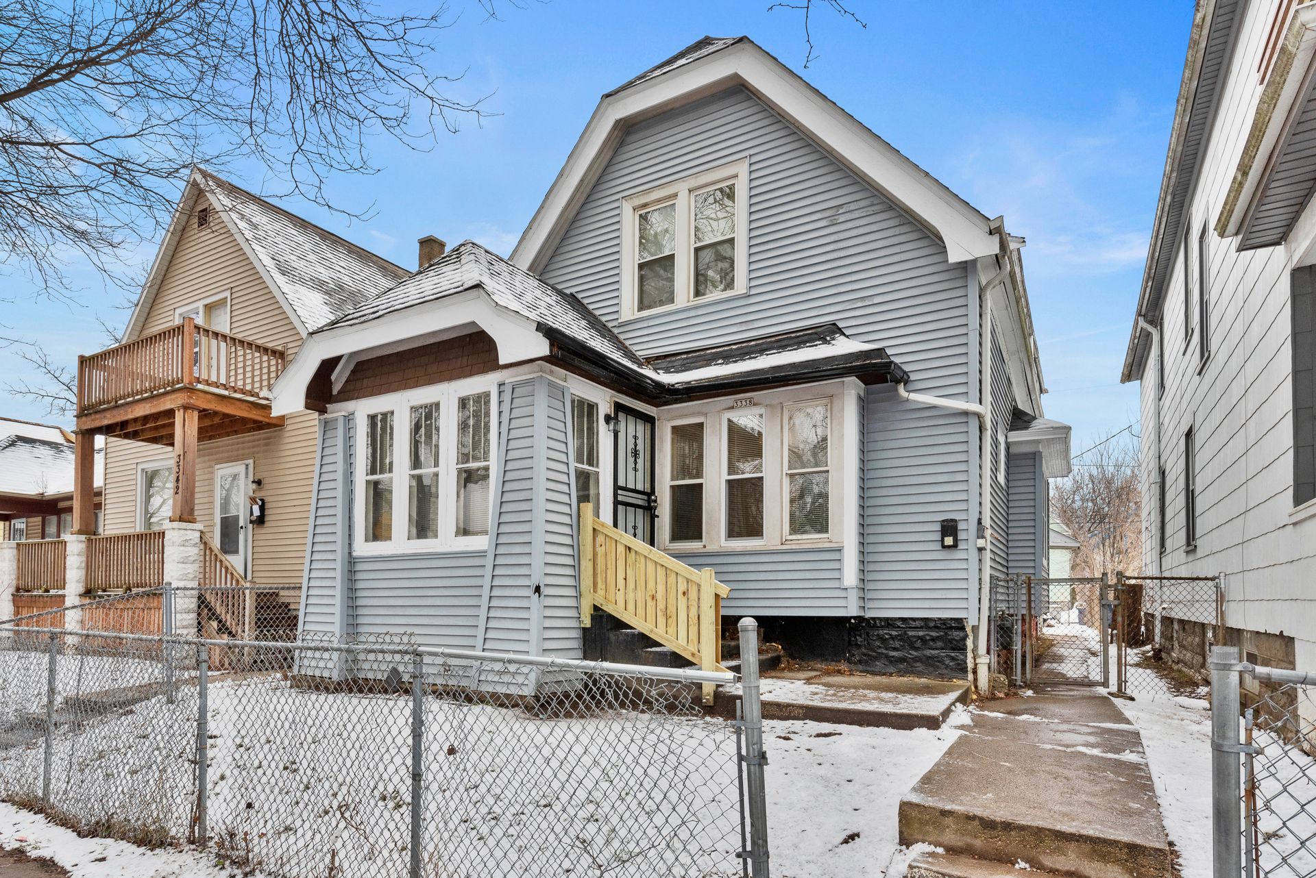 A gray house with a yellow staircase is surrounded by snow and a chain link fence.