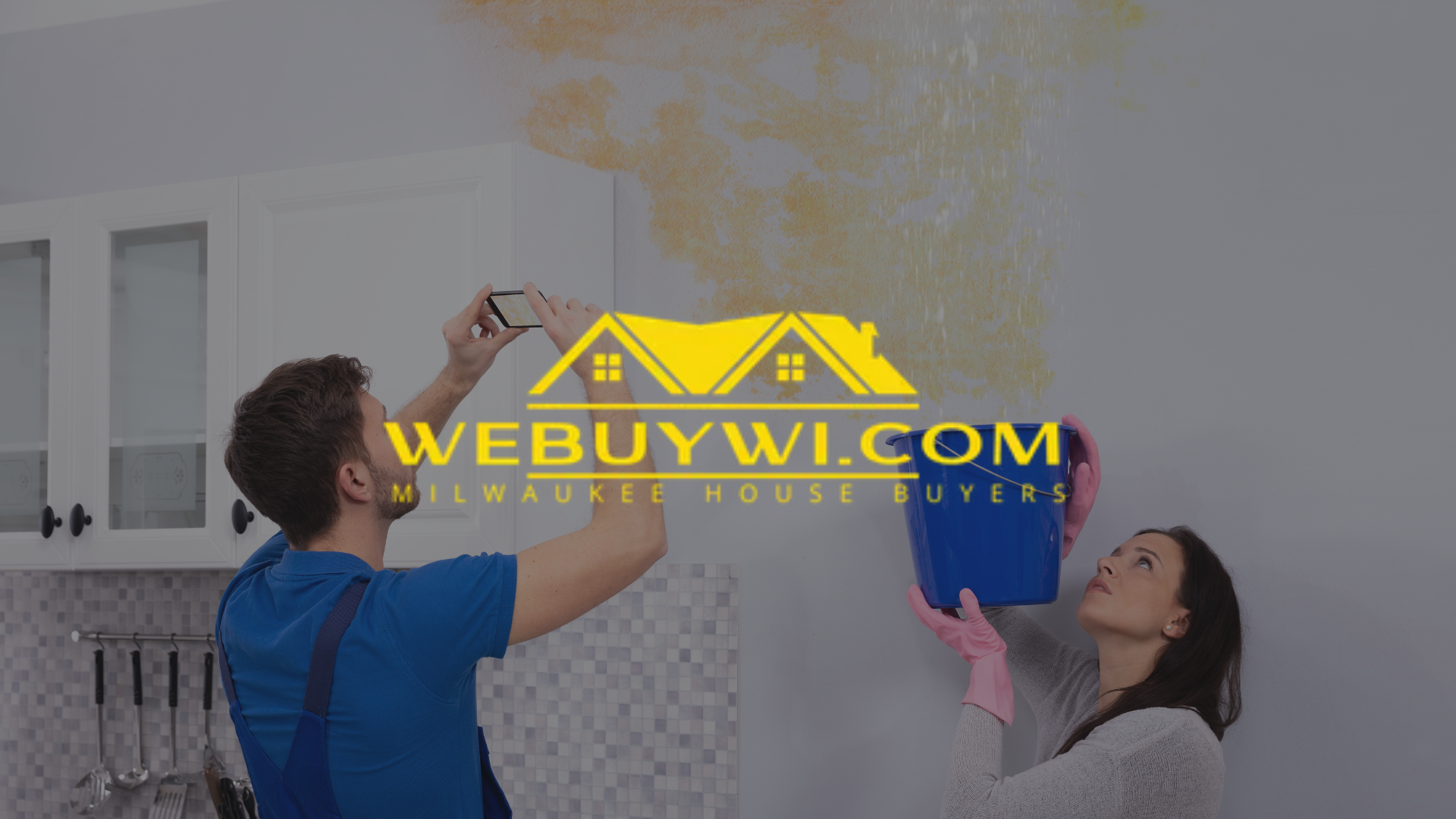 Couple in kitchen dealing with ceiling leak. Man photographs it, woman holds bucket.