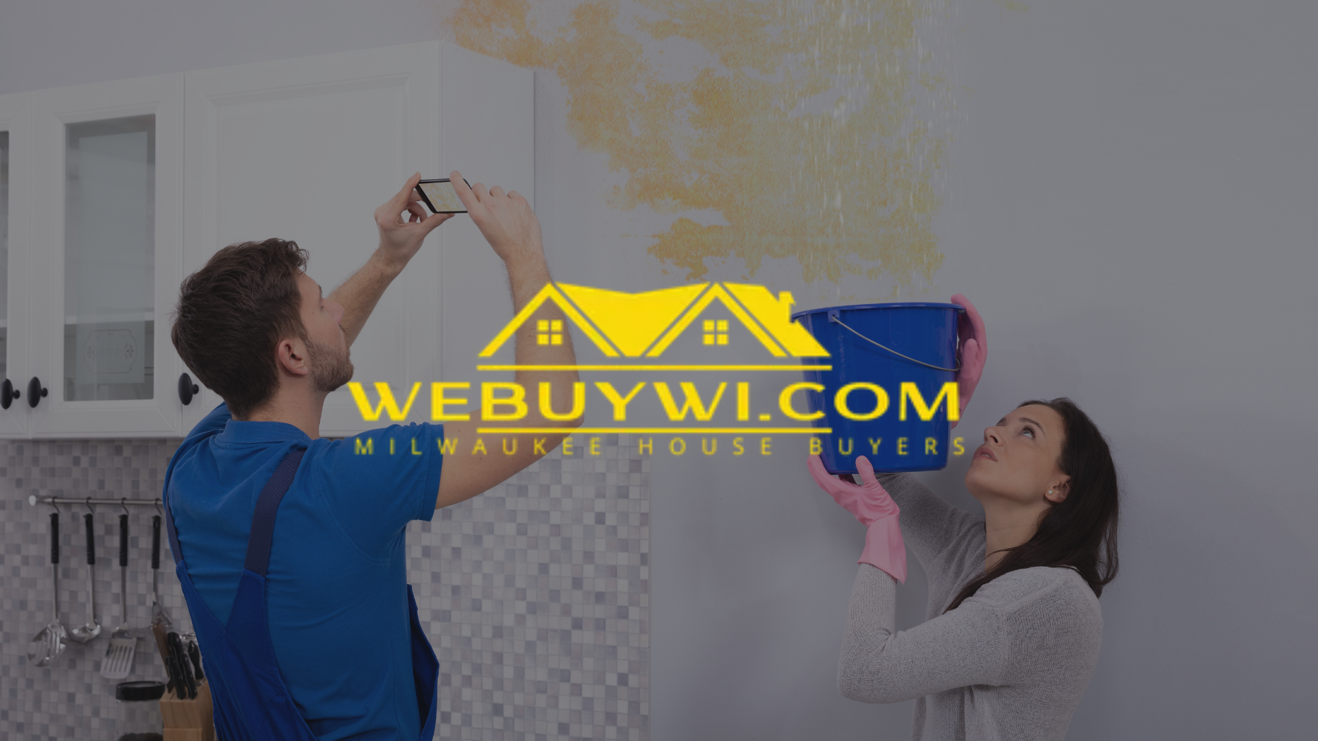 A couple inspects water damage on a kitchen wall. A man photographs the damage while a woman holds a bucket. The website logo 