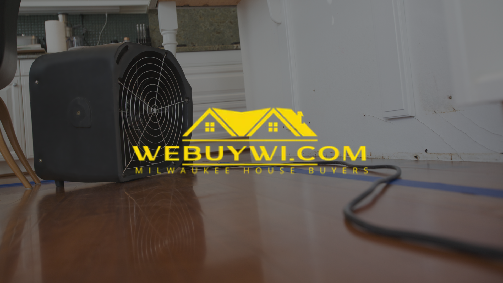 A black industrial fan is drying a shiny, wooden floor, likely after water damage. A logo for WeBuyWI is on the floor.