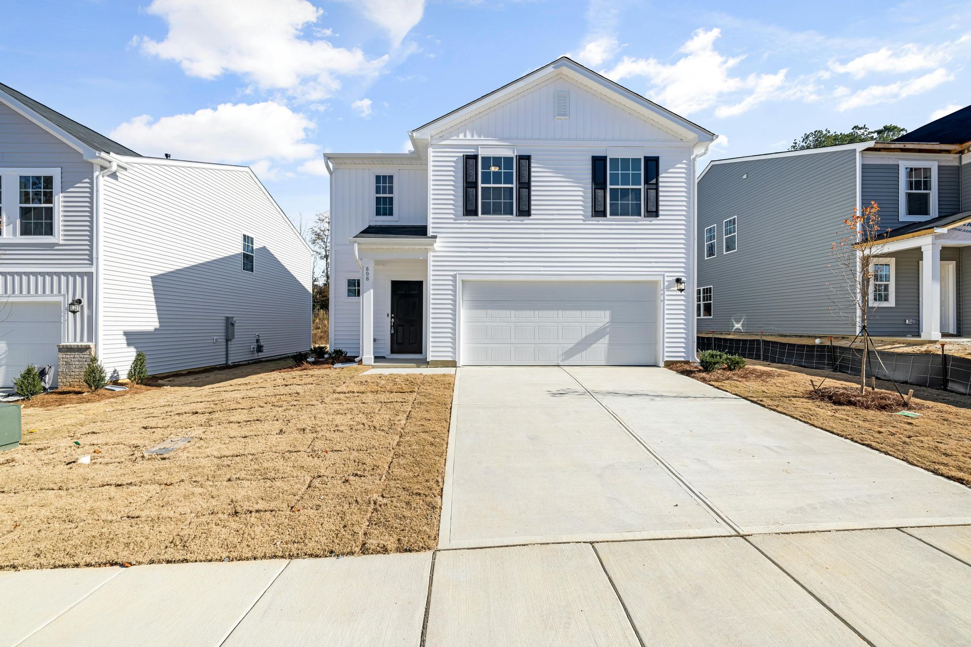 White two-story house with black shutters and a driveway. Adjacent houses under construction. Blue sky.