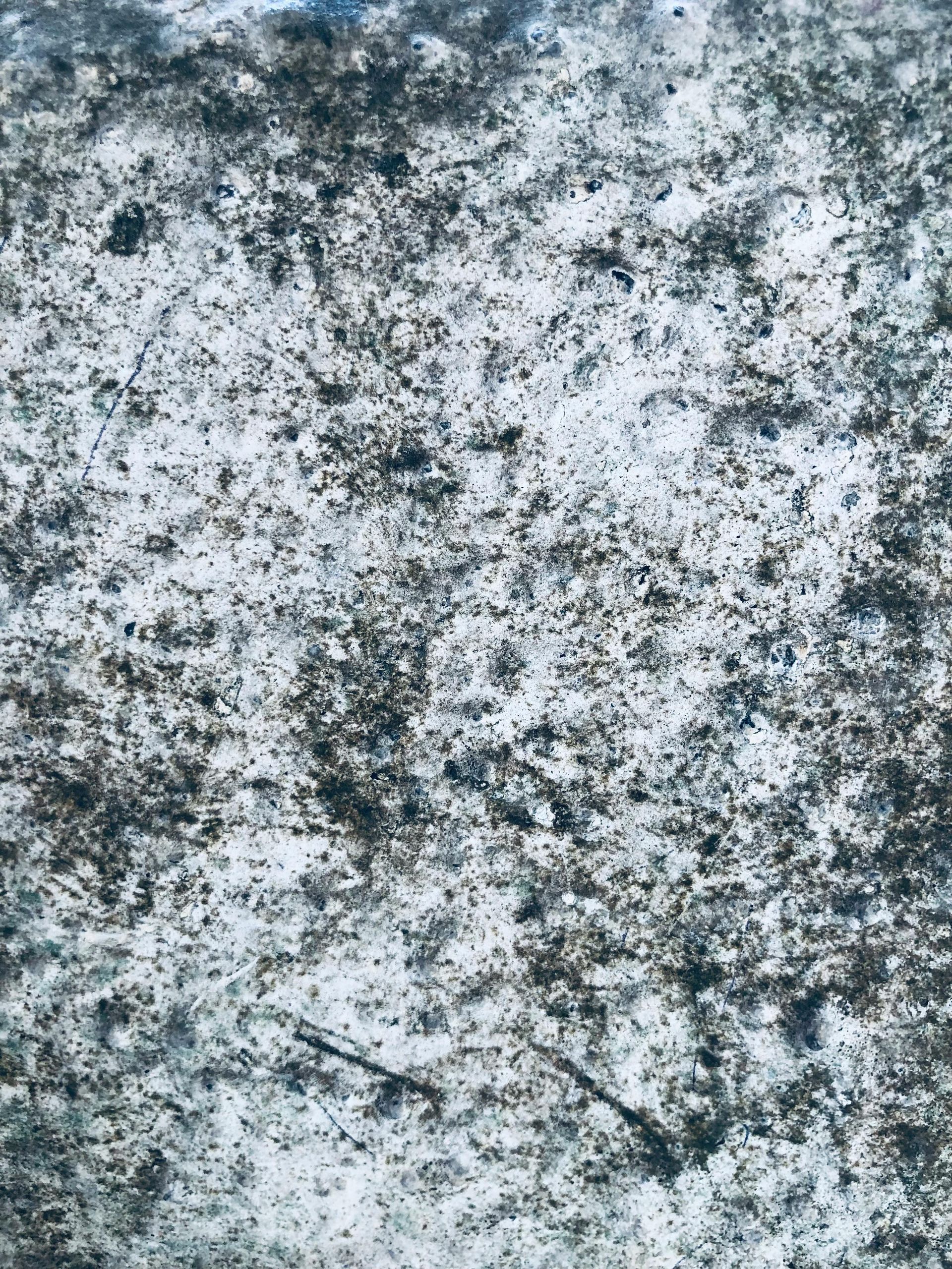 A textured, mottled surface featuring a mix of grey, black, and white tones, resembling rough concrete or weathered stone.