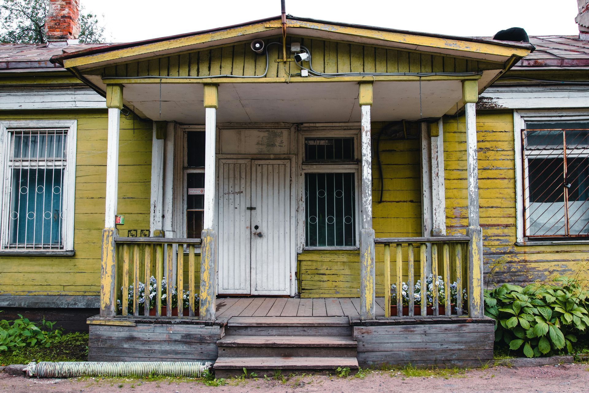 A weathered, yellow wooden house with a white front door, porch, and windows featuring iron security bars.
