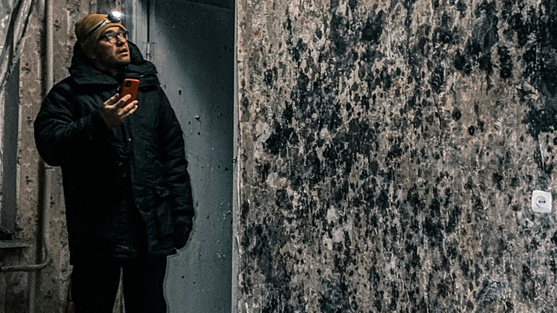 Person in a dark coat stands in a narrow alley, looking up beside a textured wall.