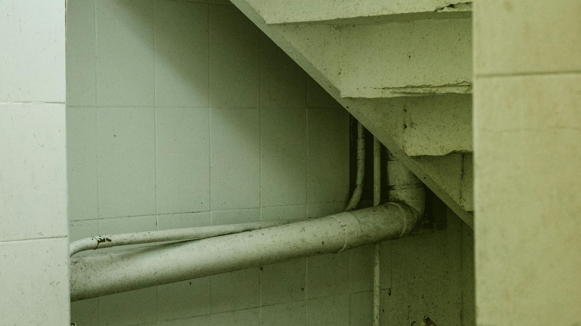 White pipe runs along a pale green wall beneath a concrete staircase.