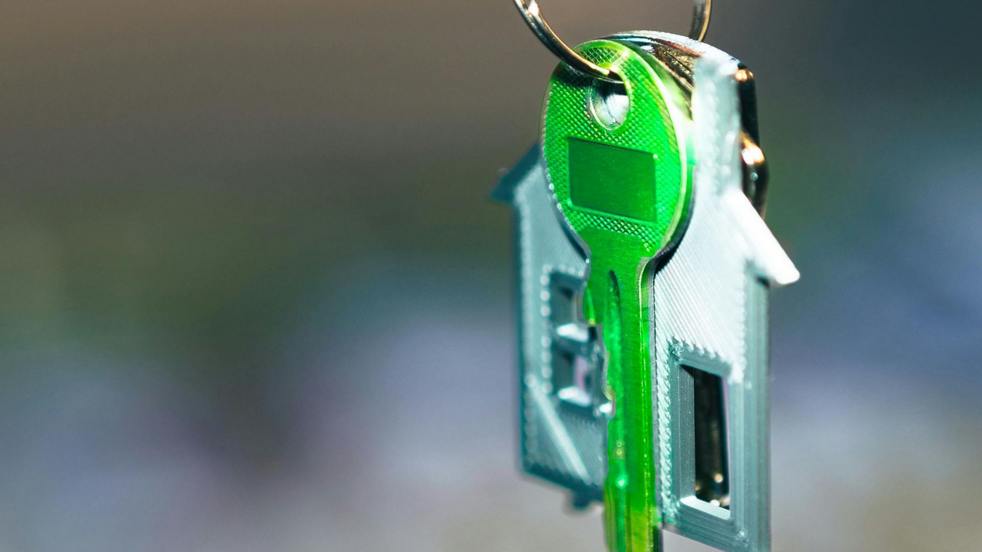 A bright green key attached to a small, silver house-shaped keychain hanging against a soft, blurred background.