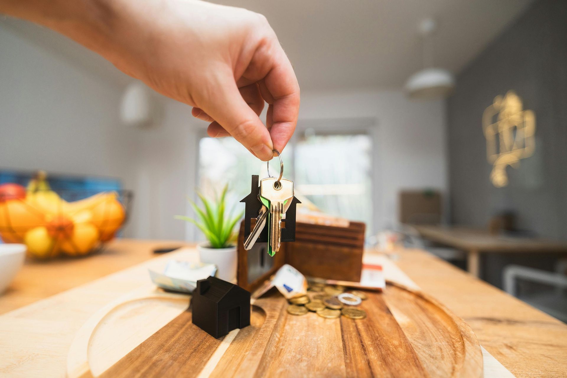 Hand placing a small model house on a wooden table with coins and a plant in the background