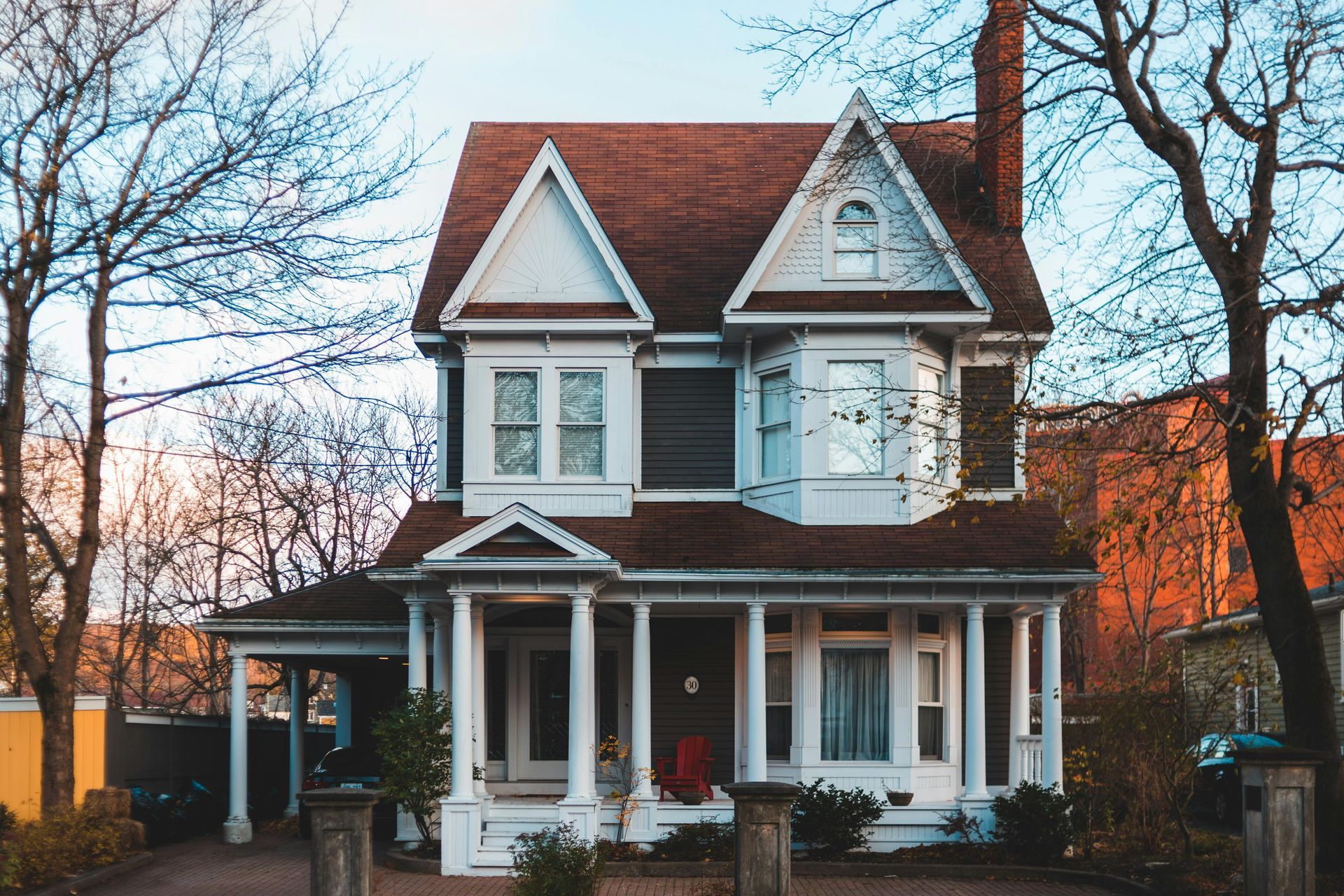 A two-story Victorian house with white pillars on the front porch, brown shingled roof, and trees on both sides.