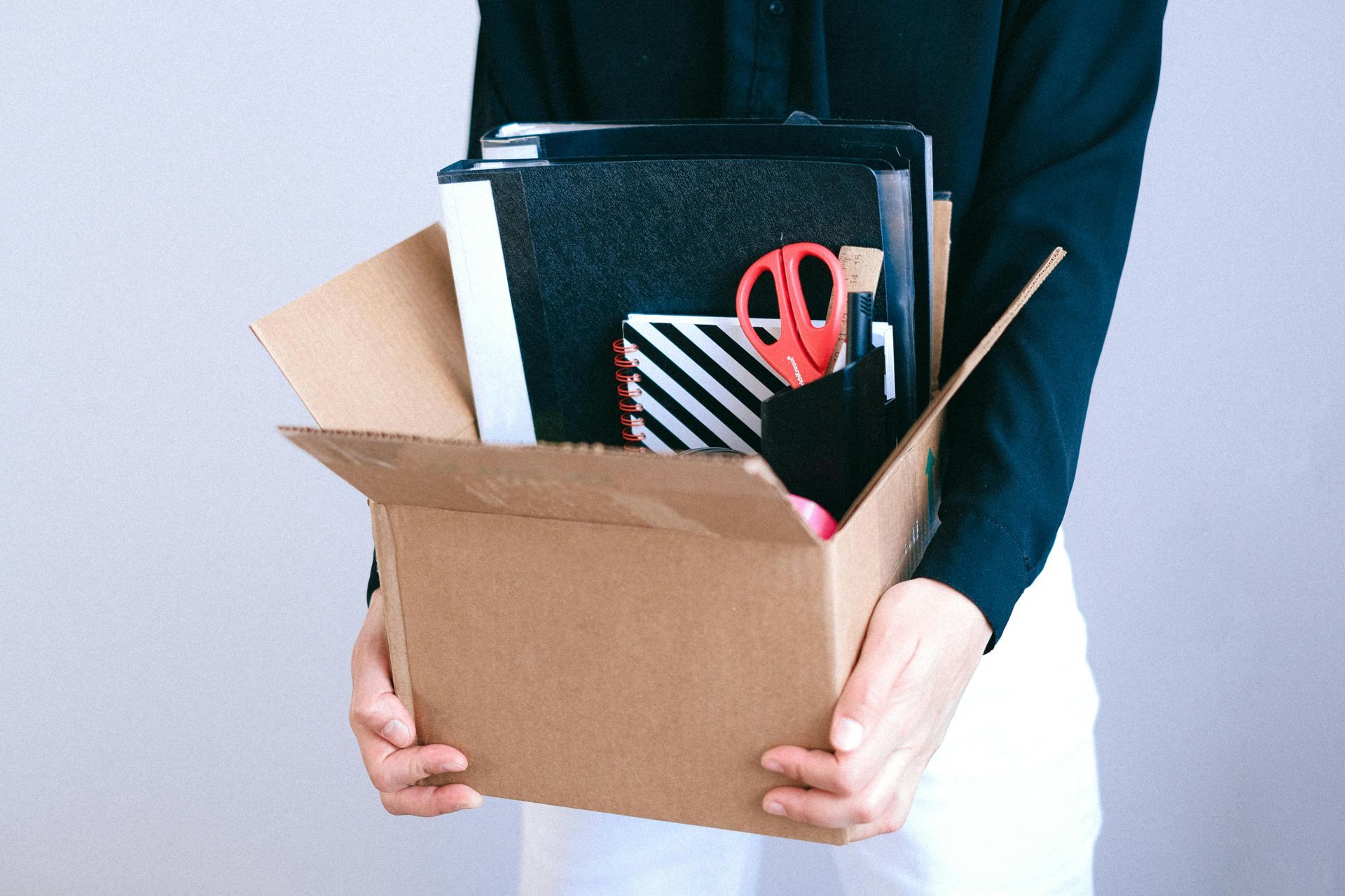 A person in a black shirt holds a cardboard box containing office supplies, including a binder, notebook, and scissors.