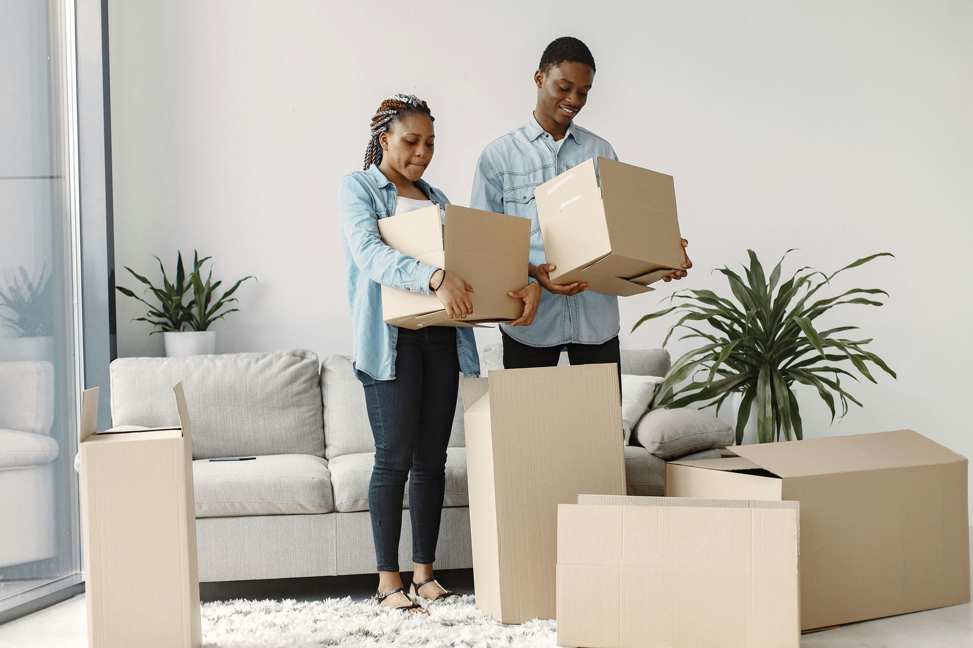 Two people in denim shirts hold cardboard boxes in a living room filled with packed boxes for moving.