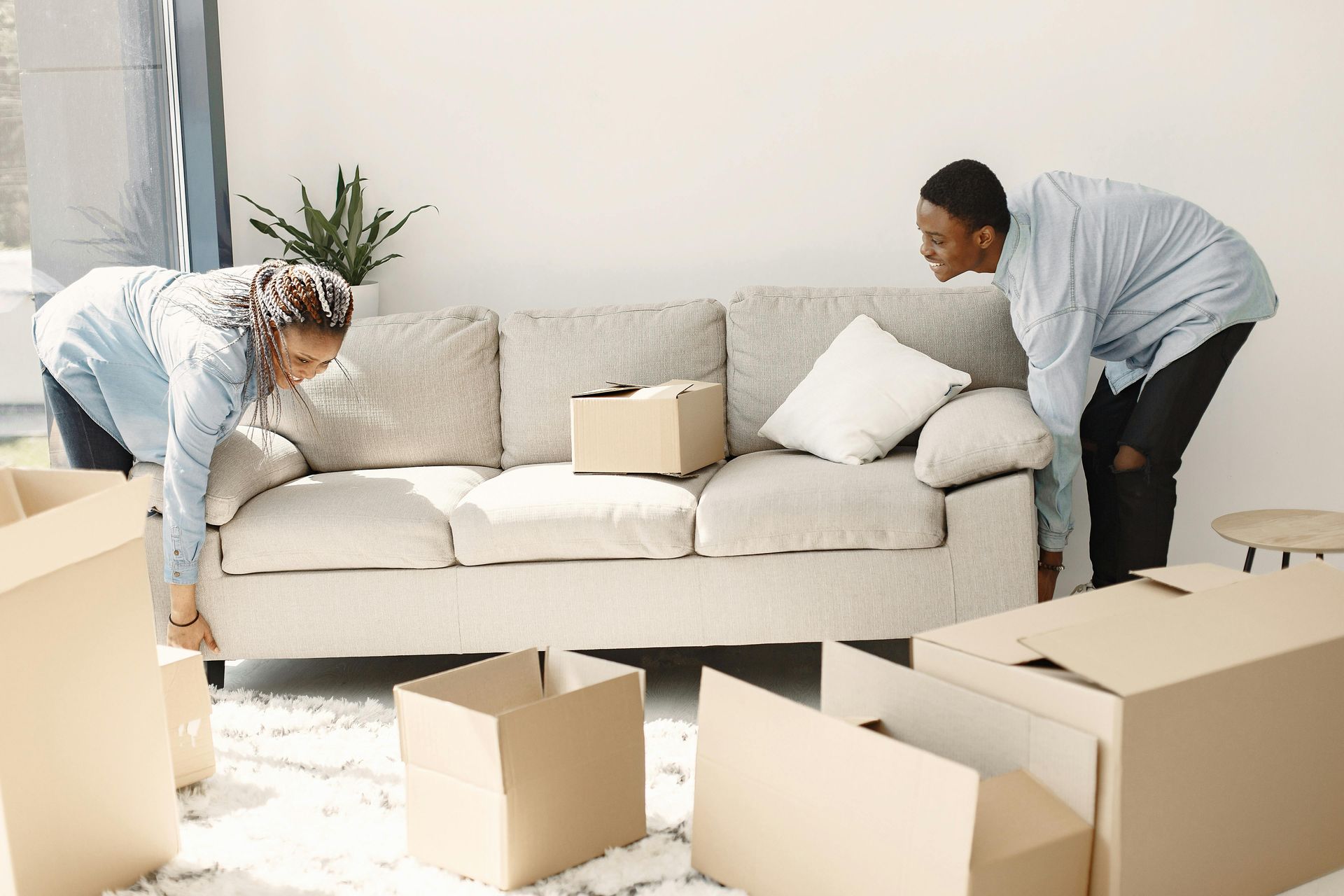 Two people move a light-colored sofa in a bright, modern living room filled with cardboard moving boxes.