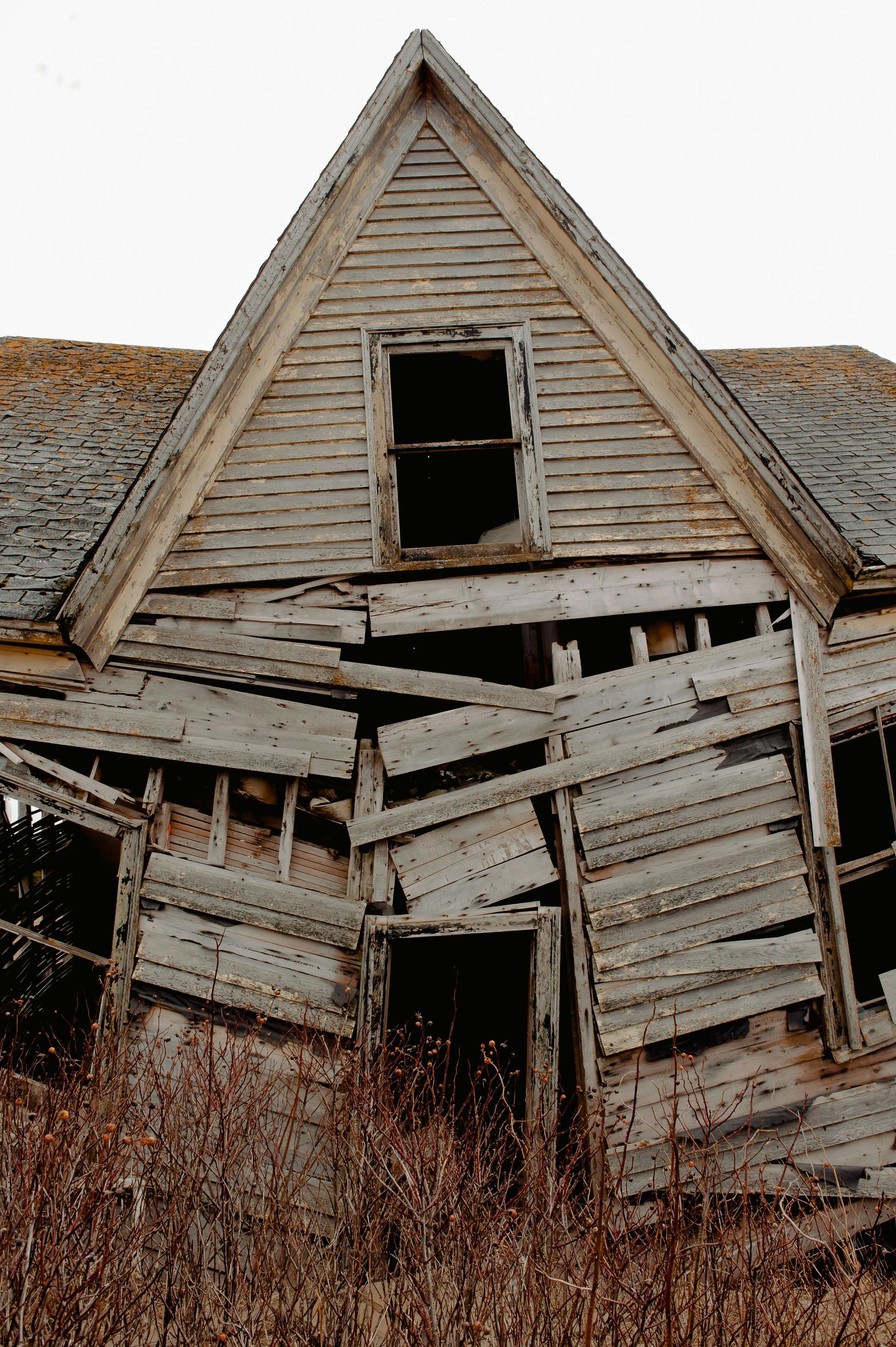 Dilapidated white wooden house with a steep gable roof and broken, leaning front porch.