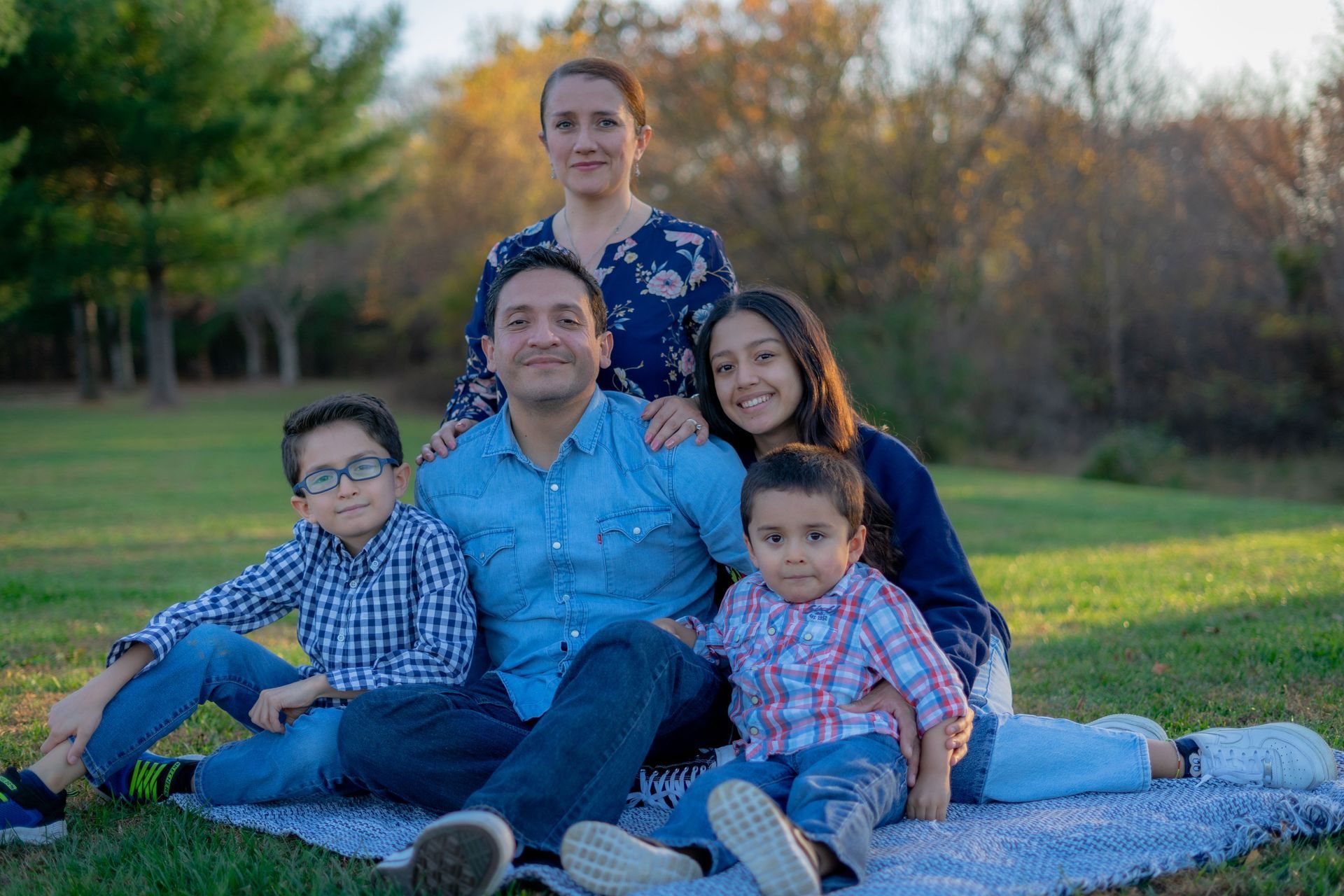 Family of five sitting on a blanket in a grassy park, with autumn trees in the background.