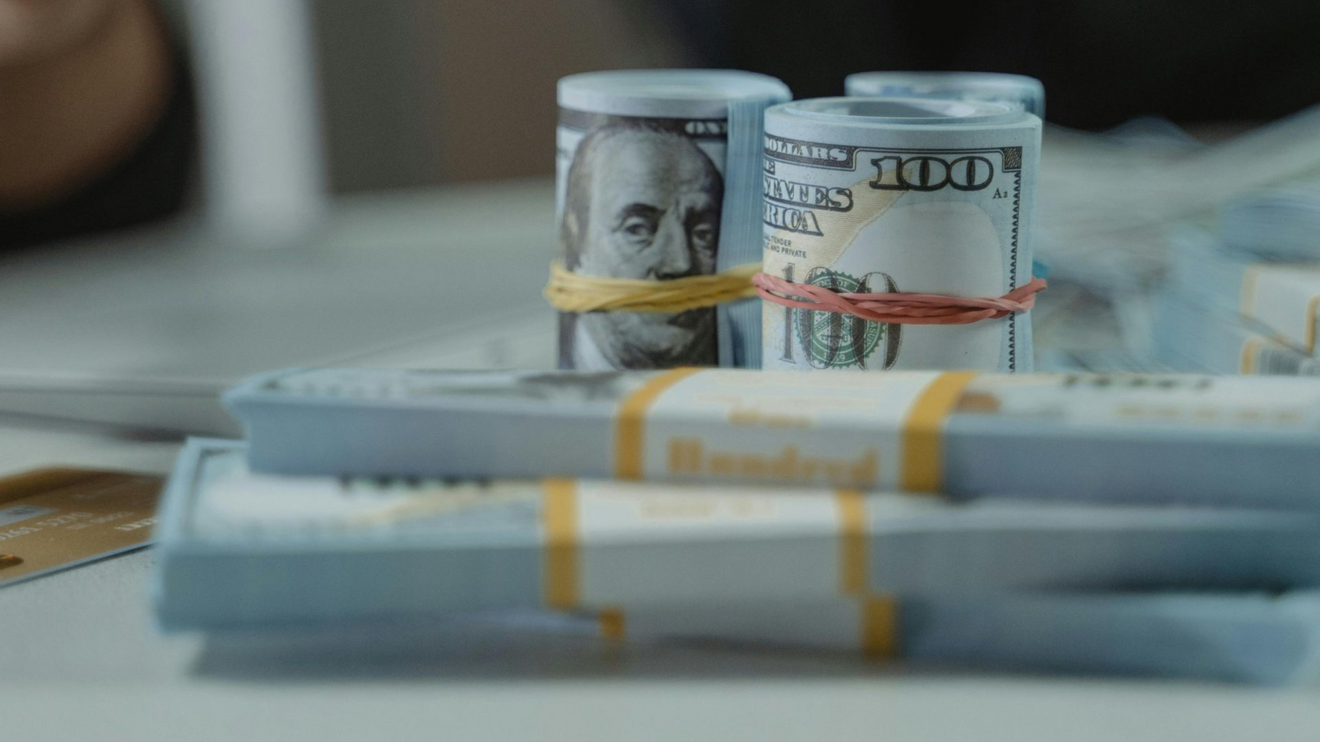 Stacks of rolled $100 bills on a table, with one bill banded in the foreground and two upright rolls behind.
