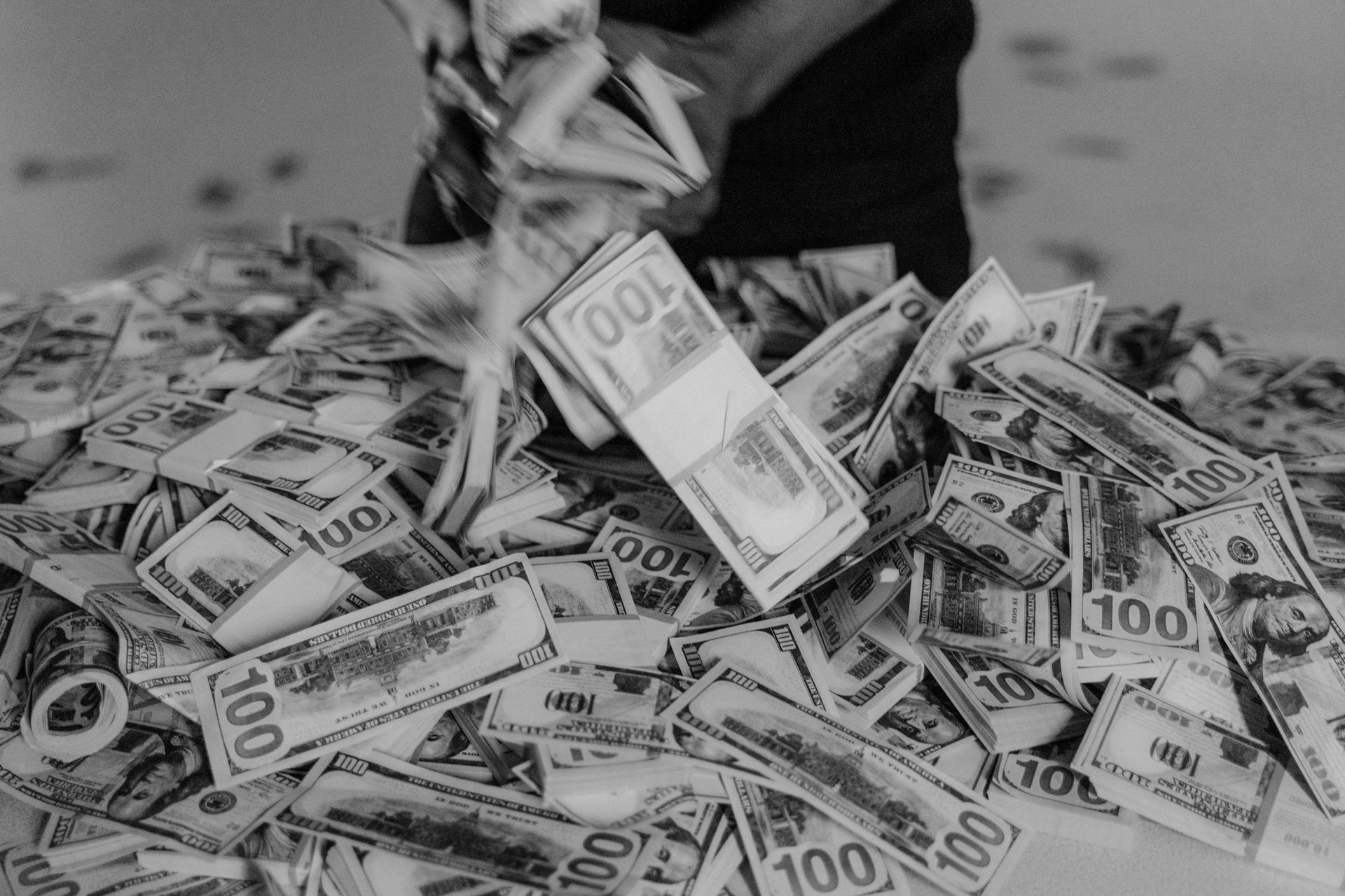 A close-up of a person's hands holding and scattering a large pile of one-hundred-dollar bills in black and white.