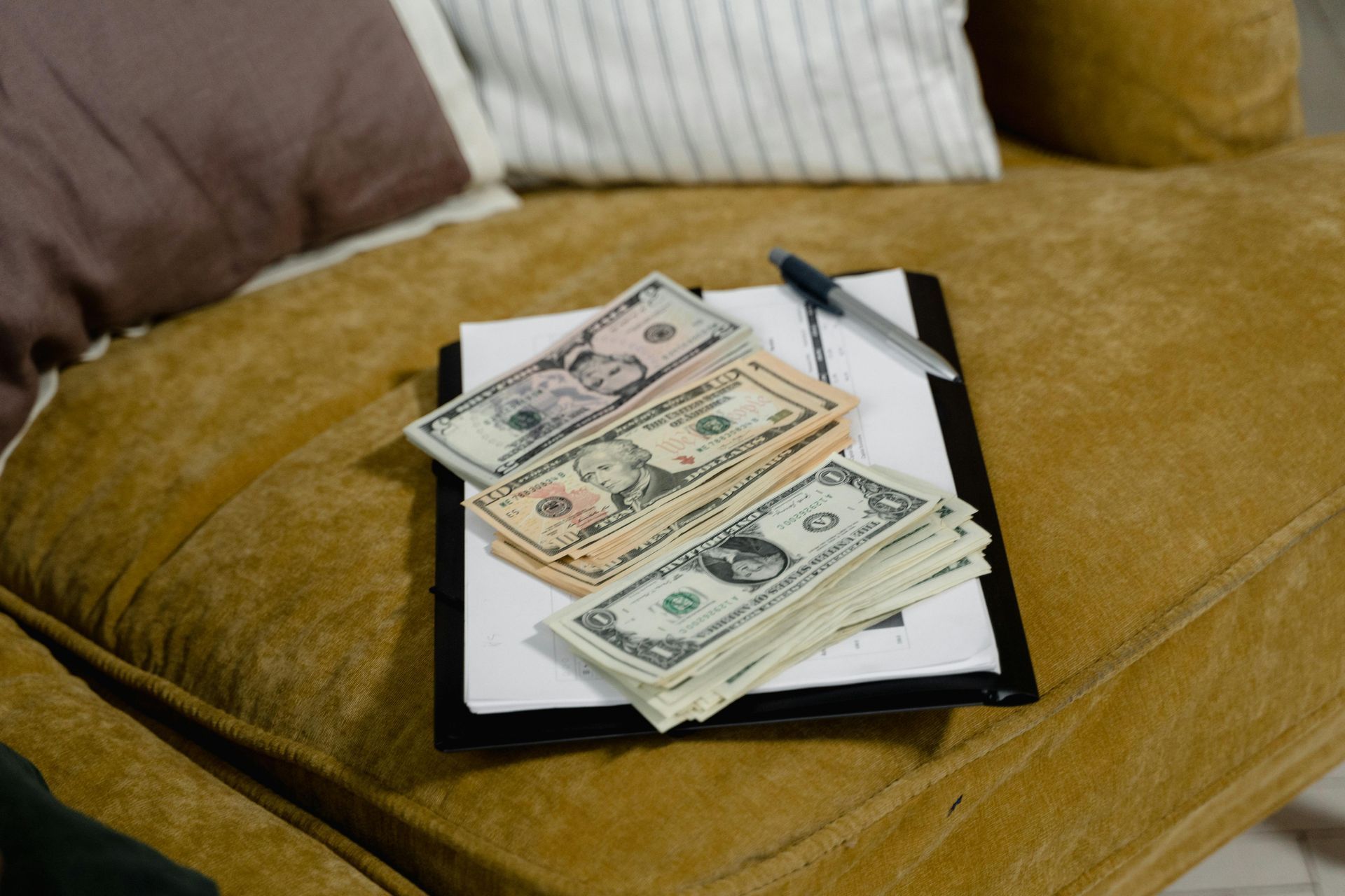 A stack of various US dollar bills rests on a clipboard with a pen, placed on a textured yellow couch.