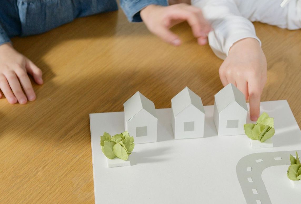 Two sets of hands arranged paper houses and small green plants on a white paper surface with a drawn road.
