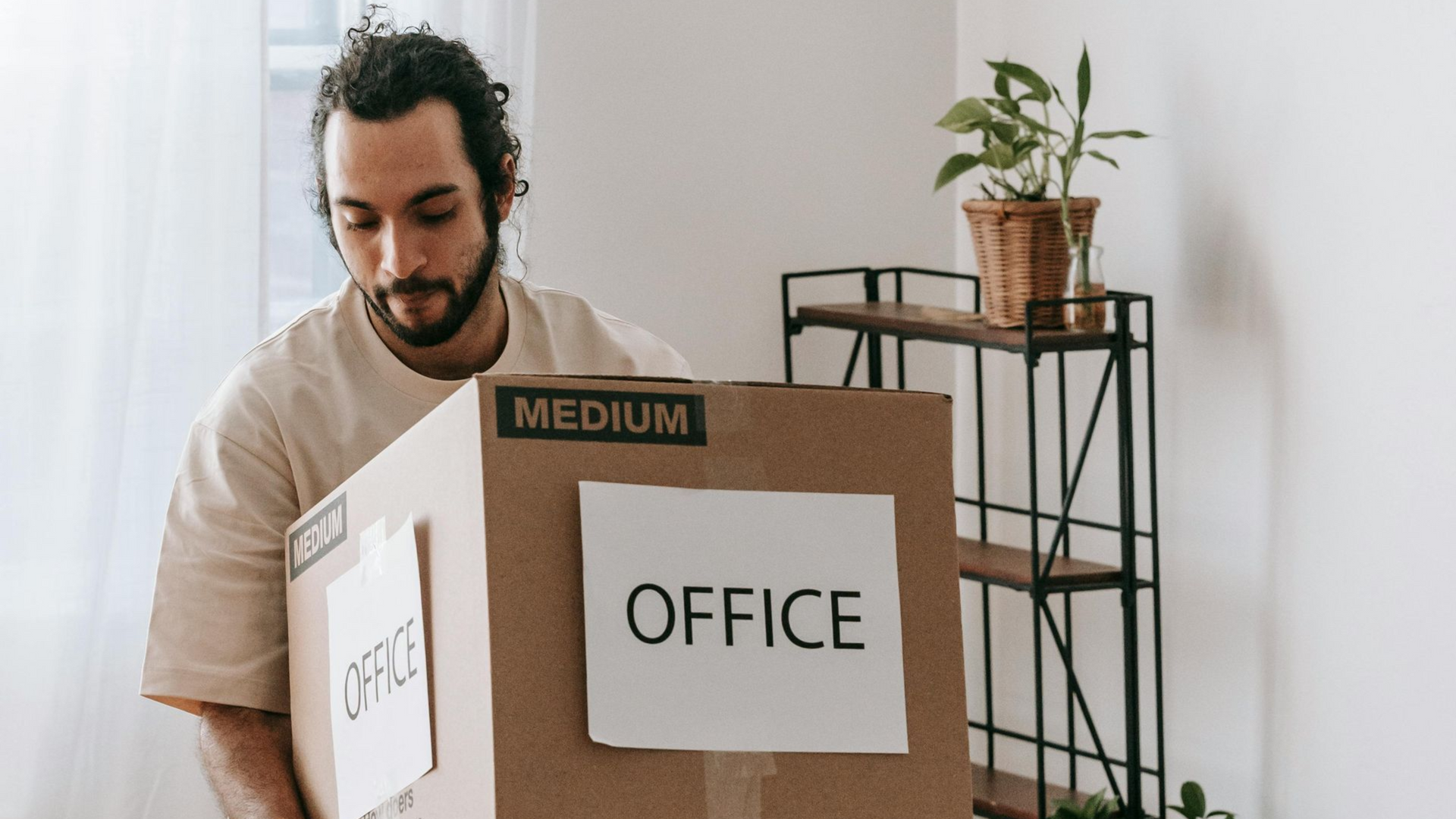 A person in a beige shirt carries a cardboard moving box labeled