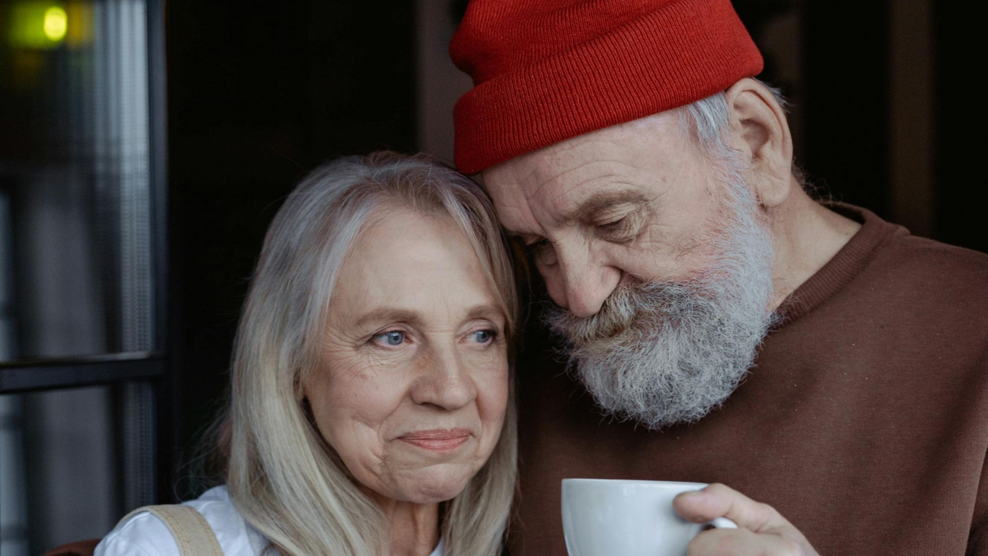 An older couple looking at each other affectionately. Man in red hat holds a white mug.