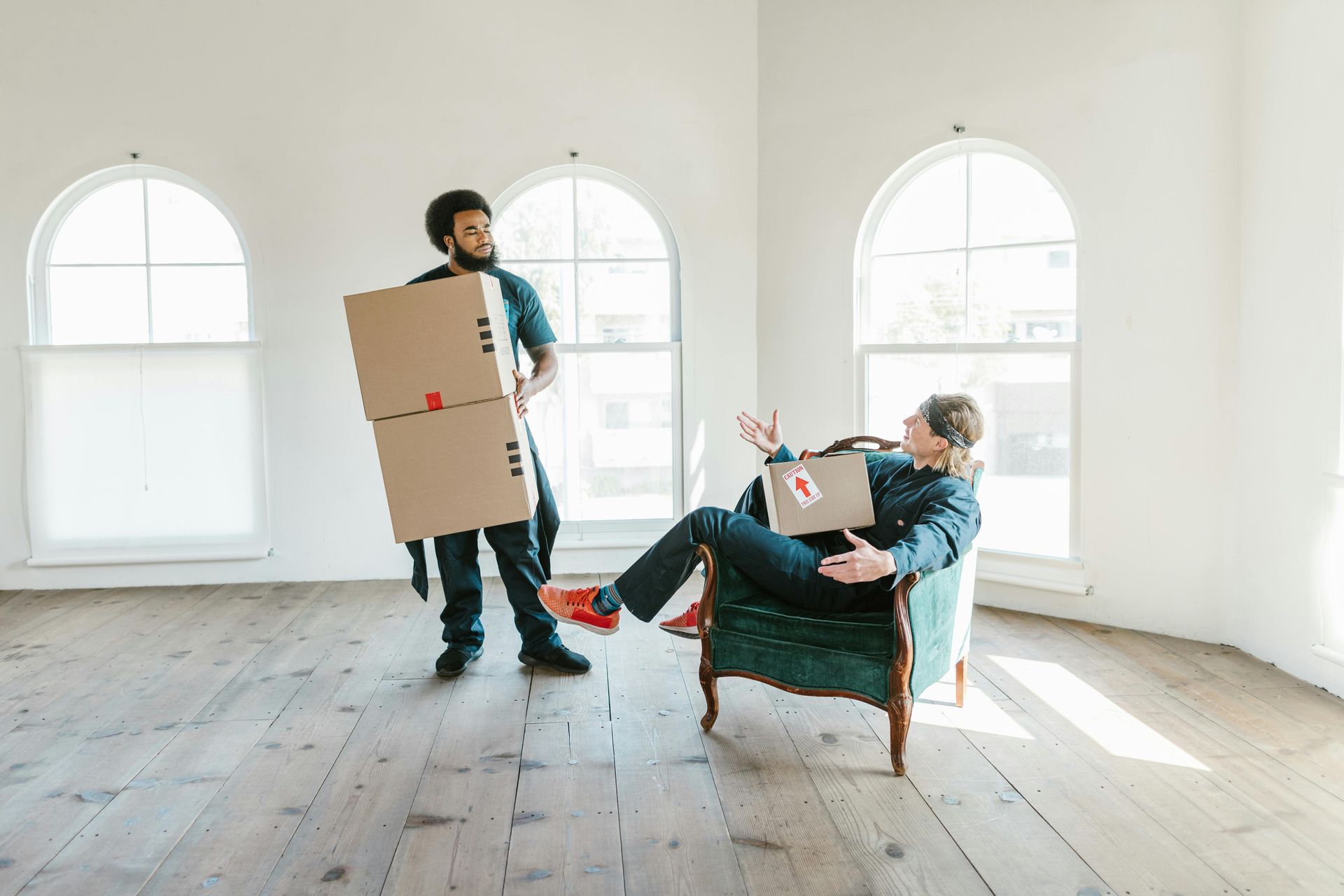 Two people in a bright, empty room with wooden floors; one carries stacked boxes while the other rests in an armchair.