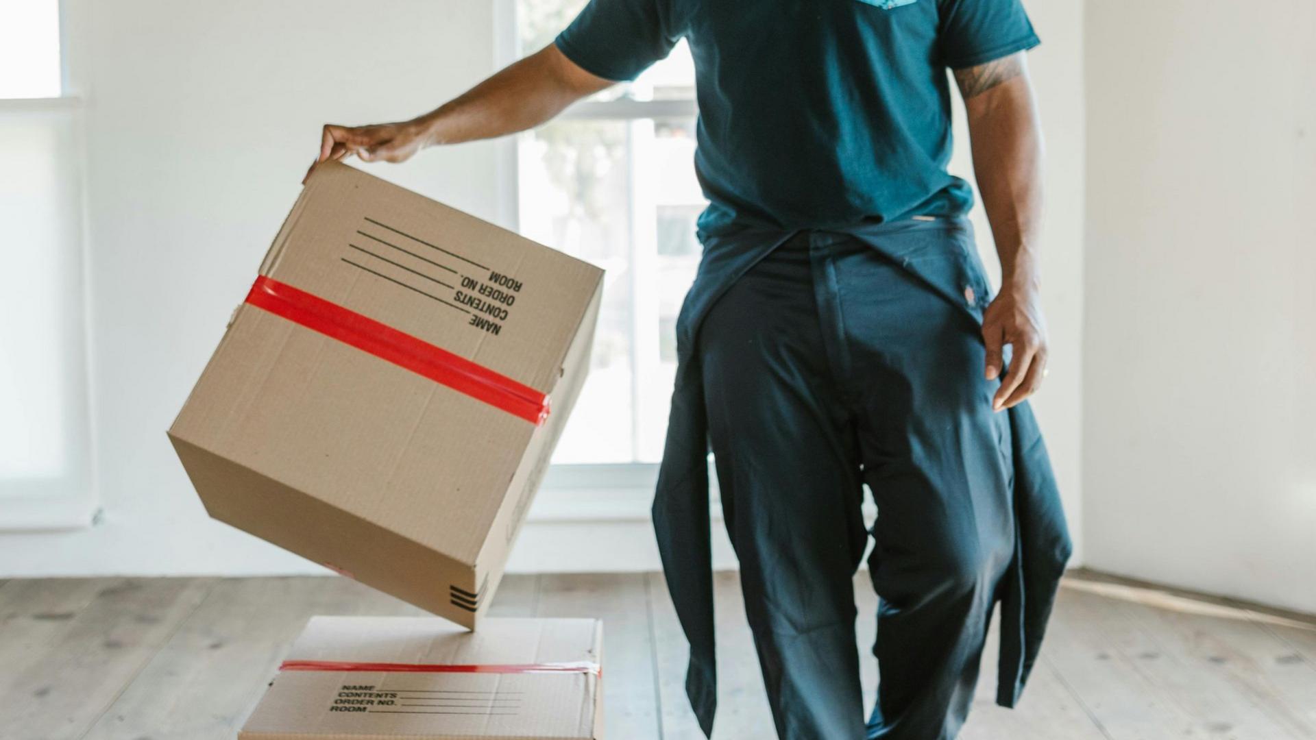 A person in dark work clothes places a brown cardboard box with a red stripe onto another box in a bright, empty room.