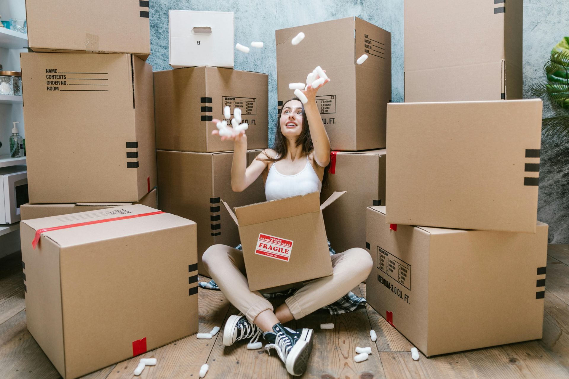 A happy person sitting on the floor surrounded by cardboard moving boxes, throwing packing peanuts into the air.