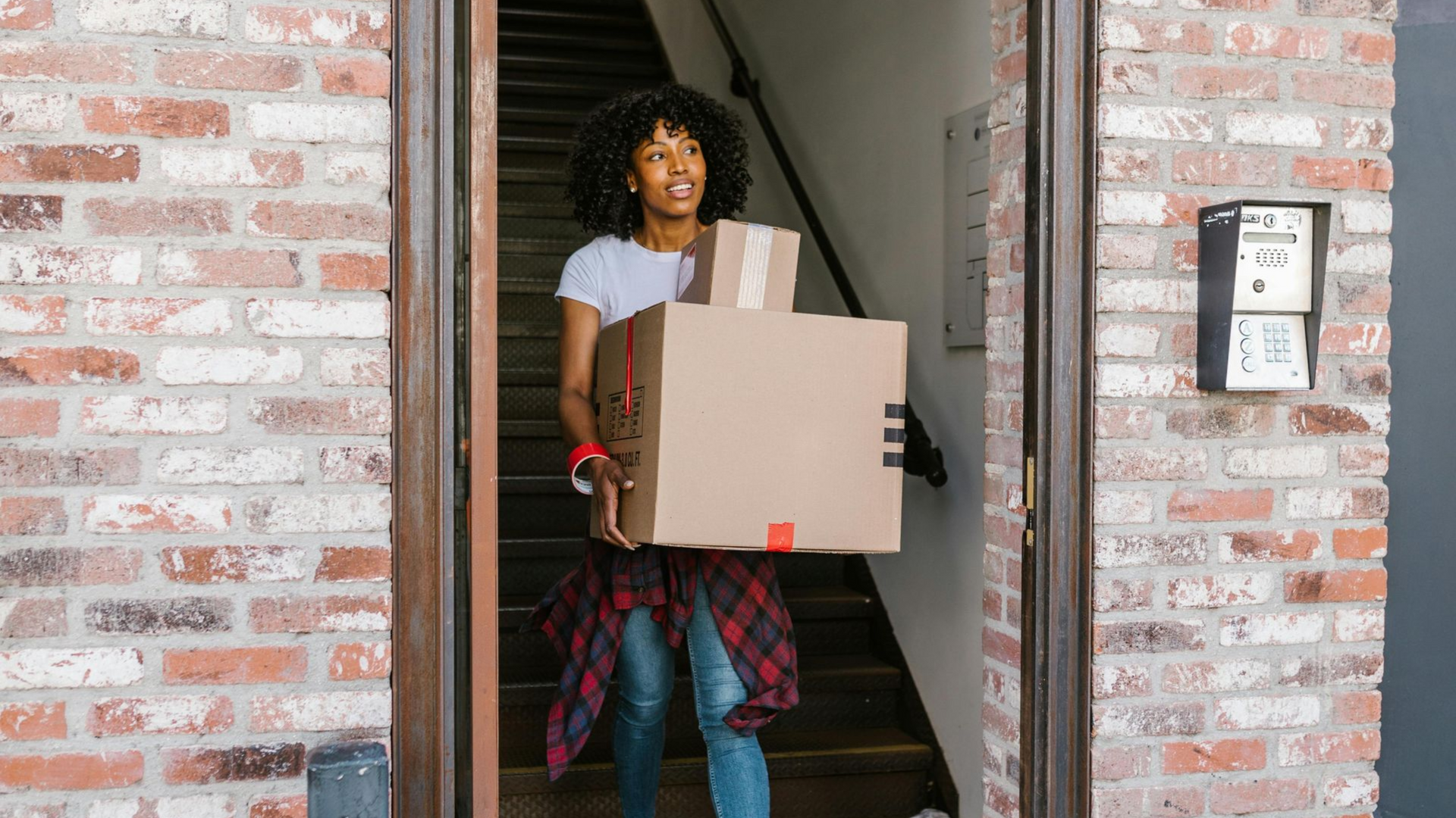 A person carries two cardboard boxes while stepping out of a doorway in a brick building.