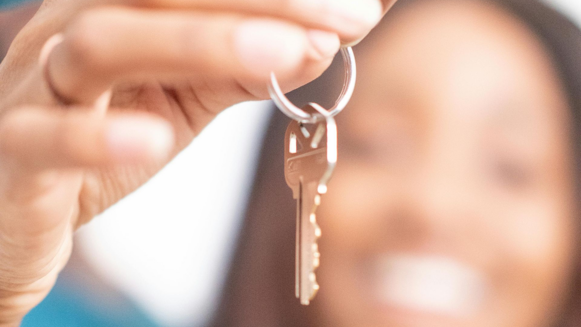 A close-up of hands holding a silver key on a keyring, with a smiling person blurred in the background.