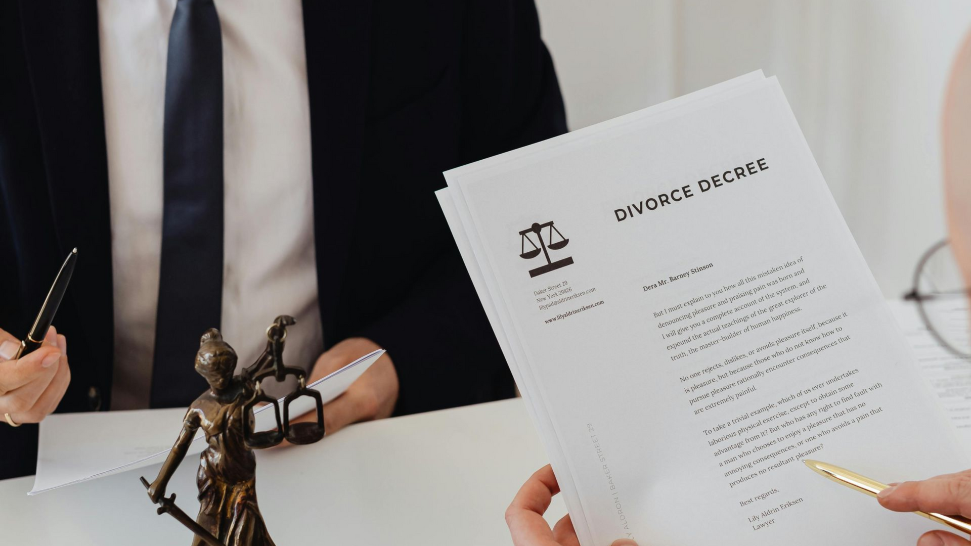 Two people in suits reviewing documents at a table, one holding a pen, a statue of Lady Justice in the foreground.