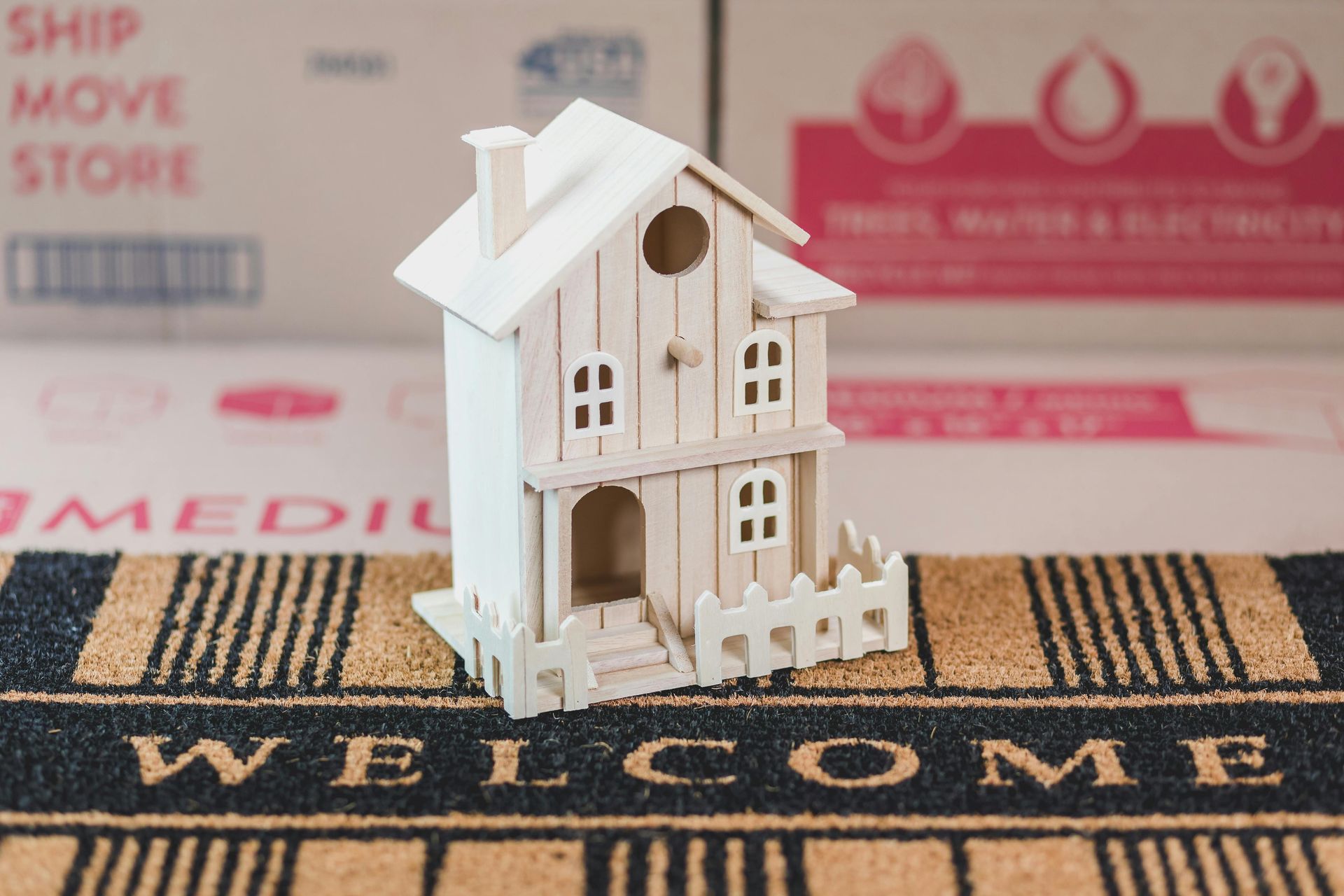 Wooden house model on a welcome mat in front of moving boxes.