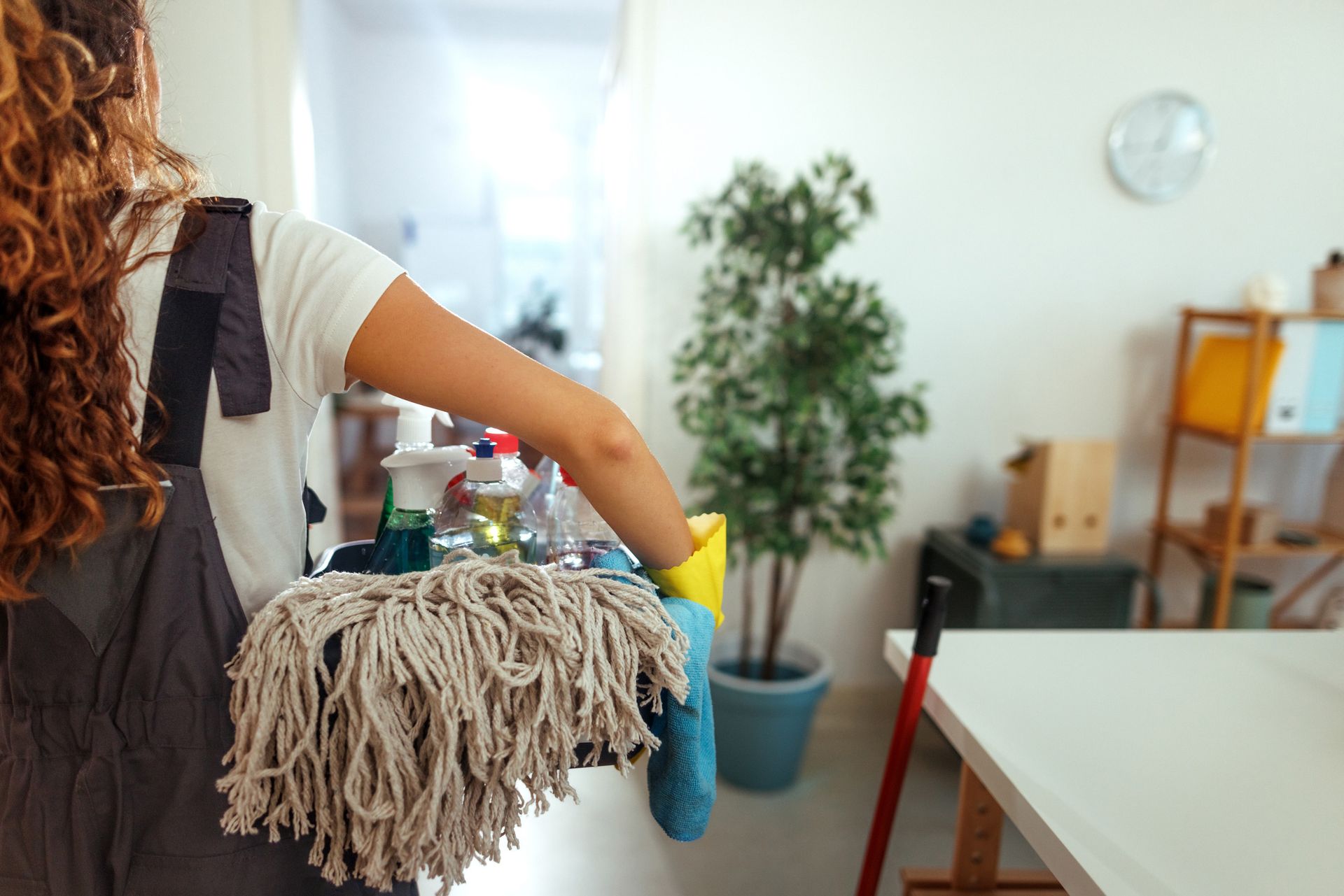 A woman is holding a mop and cleaning supplies in a living room.