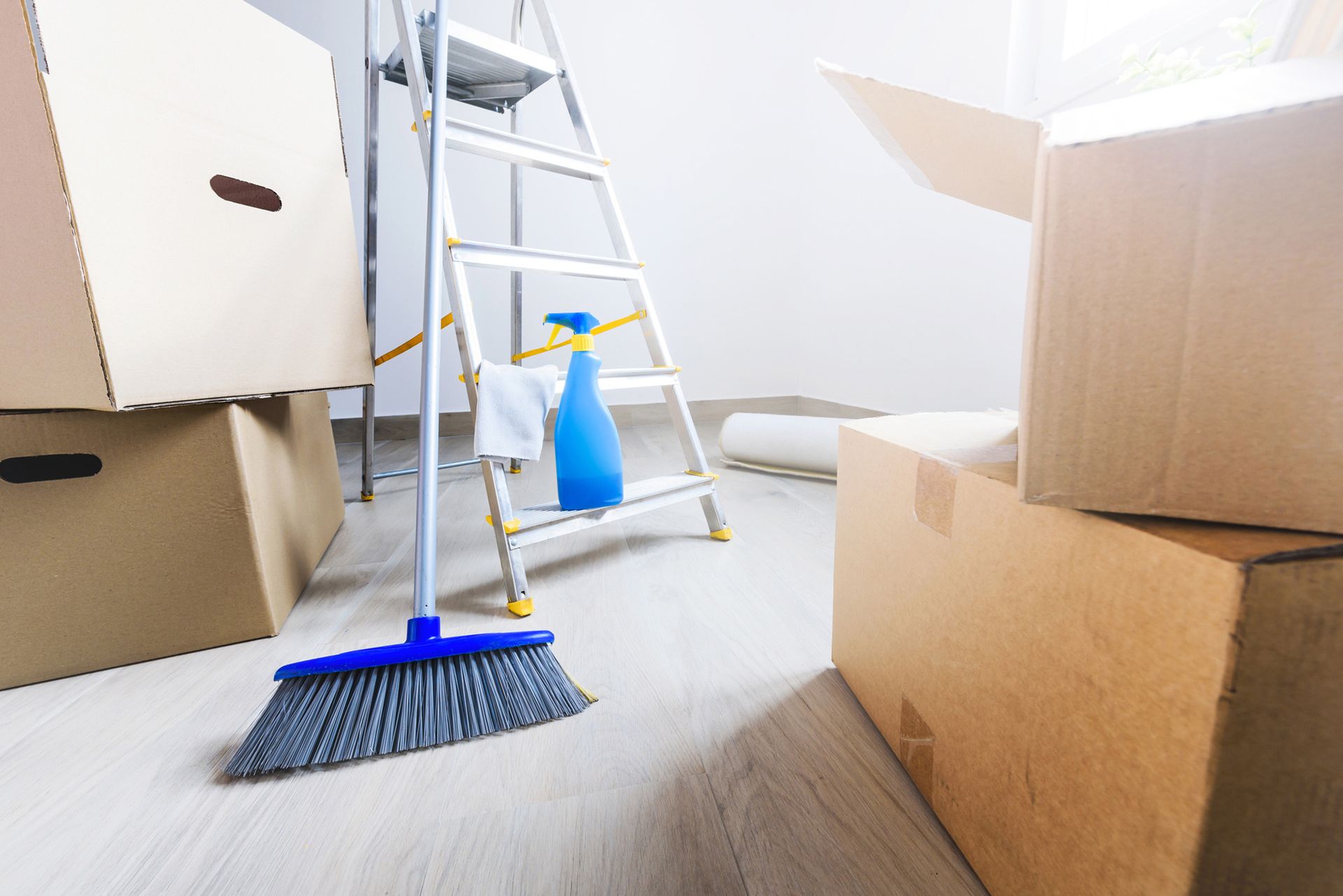 A room filled with boxes, a broom, a spray bottle and a ladder.