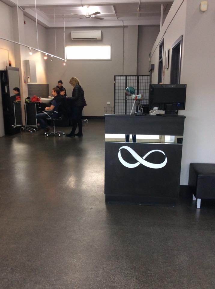 Hair salon interior with a black reception desk featuring an infinity symbol. A stylist is working on a client's hair.