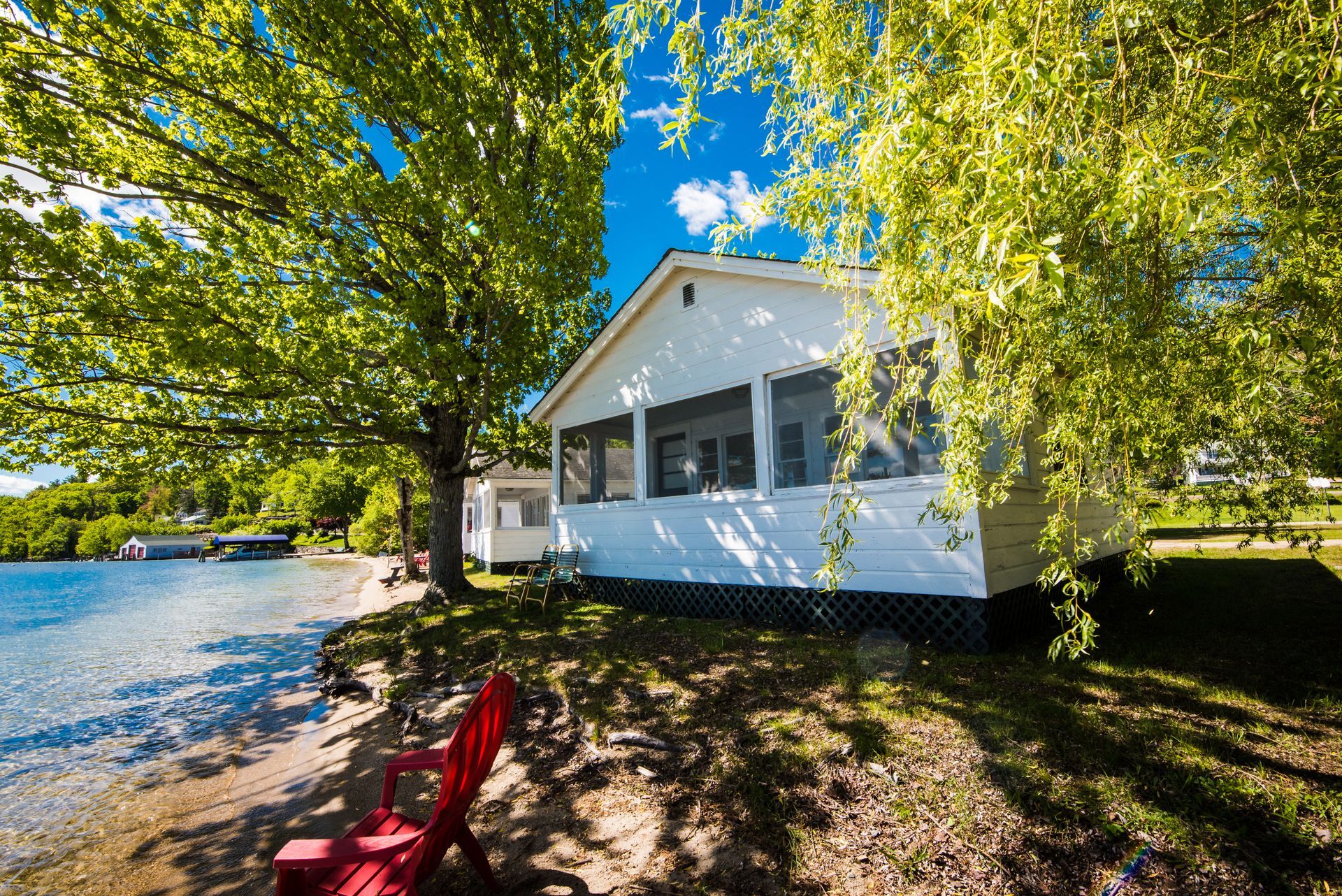 White cottage with screened porch next to a sandy beach. A red chair sits under a tree, overlooking the water.